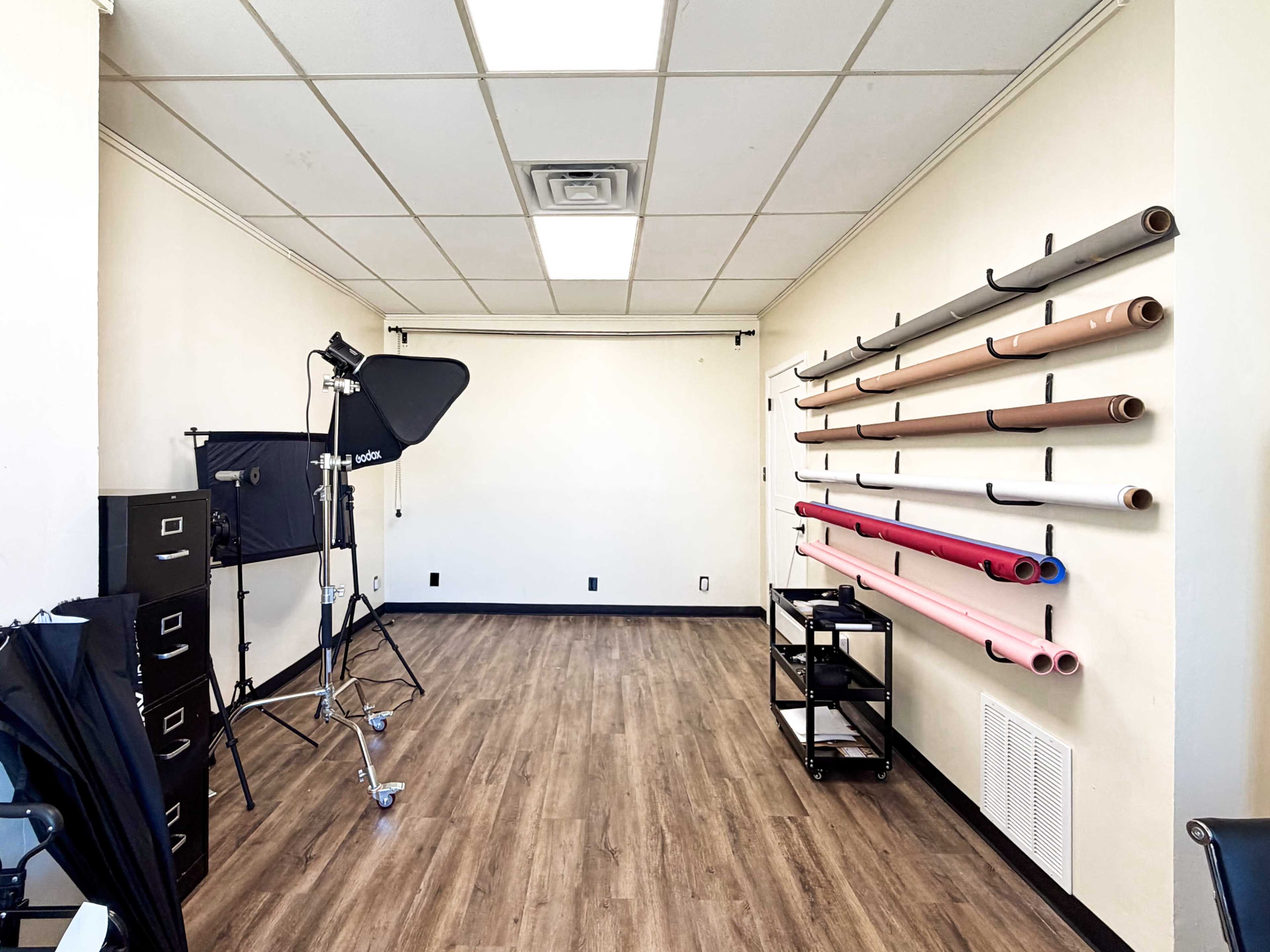 The image shows a spacious studio with a wooden floor, equipped with a backdrop stand, photography lighting, and rolls of colored paper mounted on the wall.