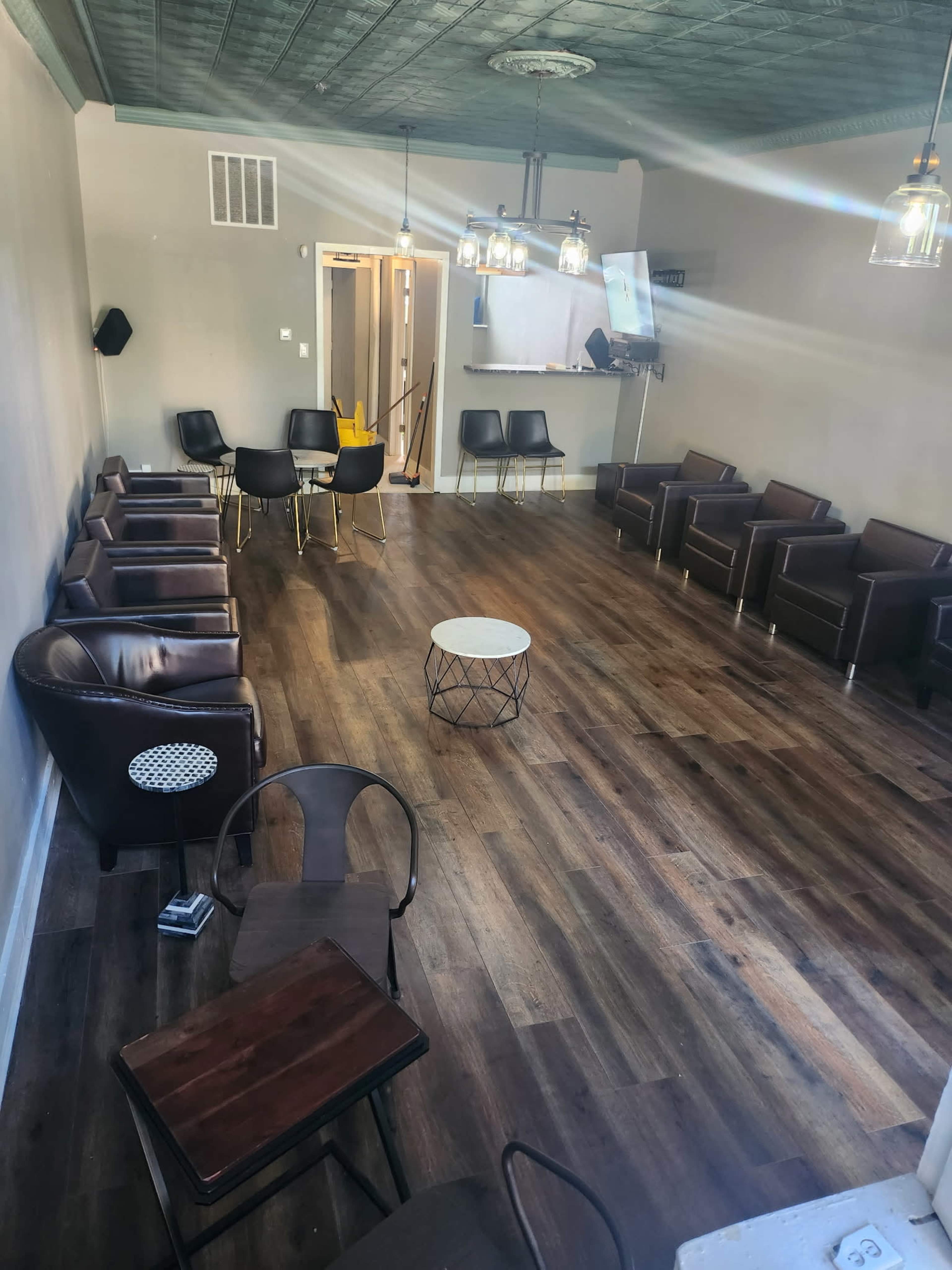 The image shows a spacious, modern lounge area with dark wood flooring, several leather chairs arranged around small tables, and a bar area in the background.