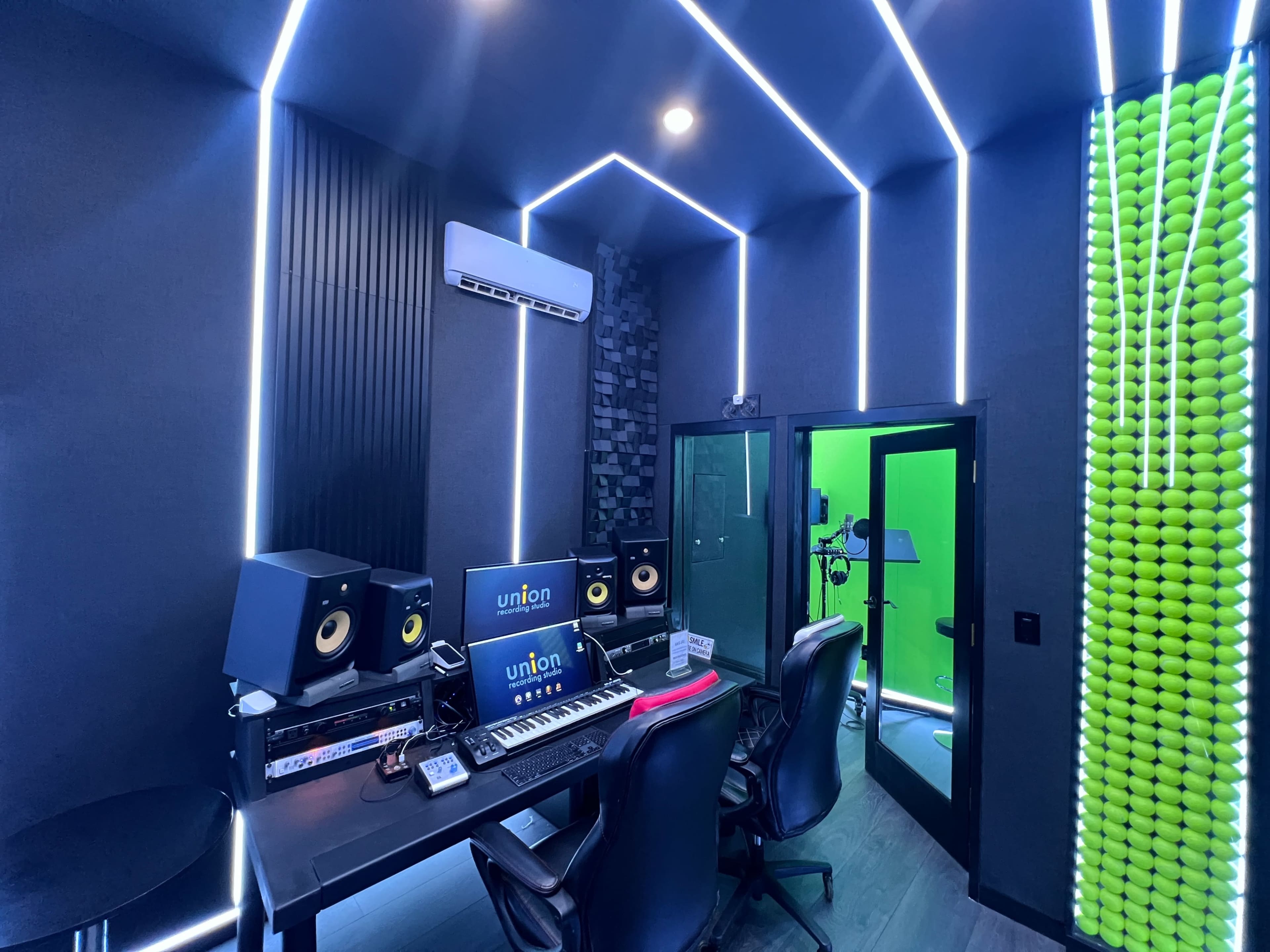 A modern music recording studio with a control desk, monitor speakers, and a vocal booth, illuminated by LED strip lighting.