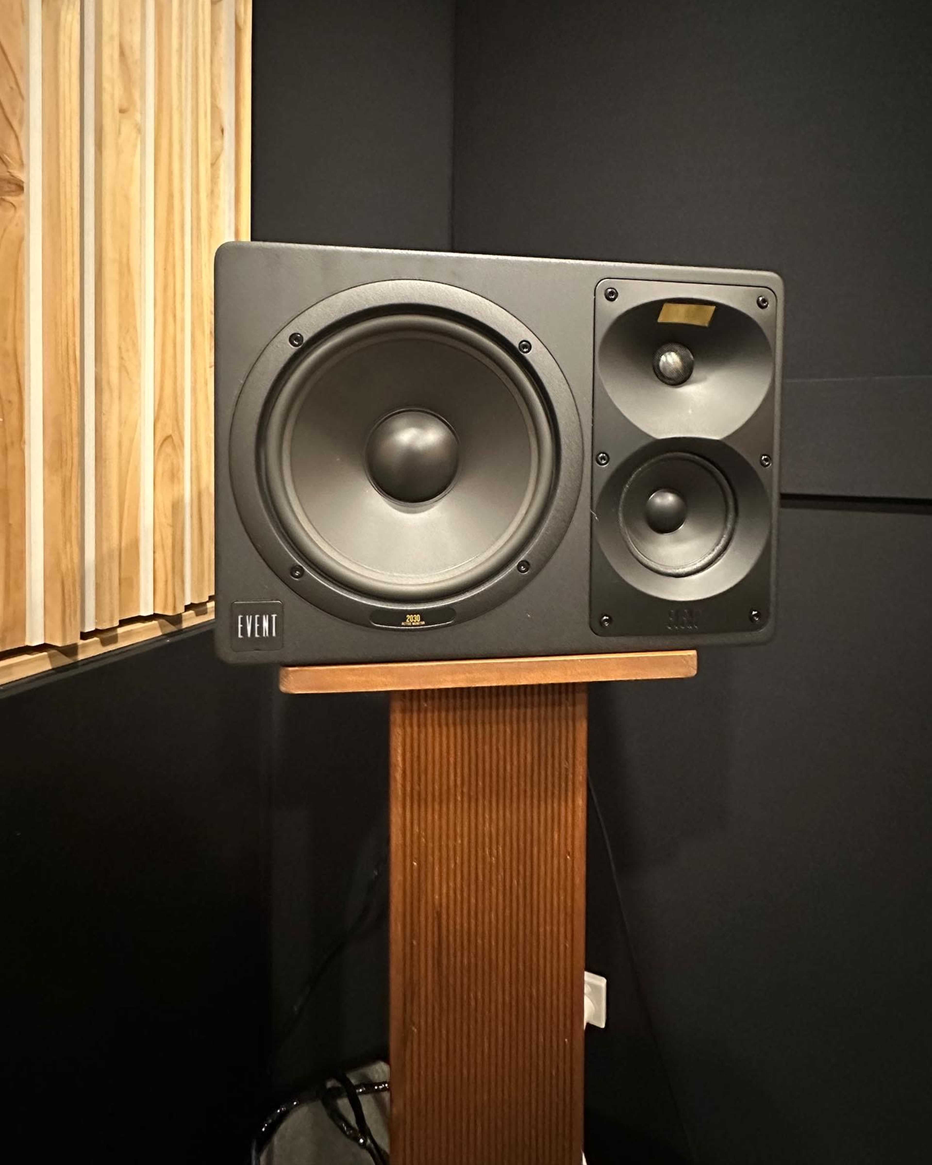 A black studio monitor speaker is placed on a wooden stand against a dark wall, with soundproof panels in the background.
