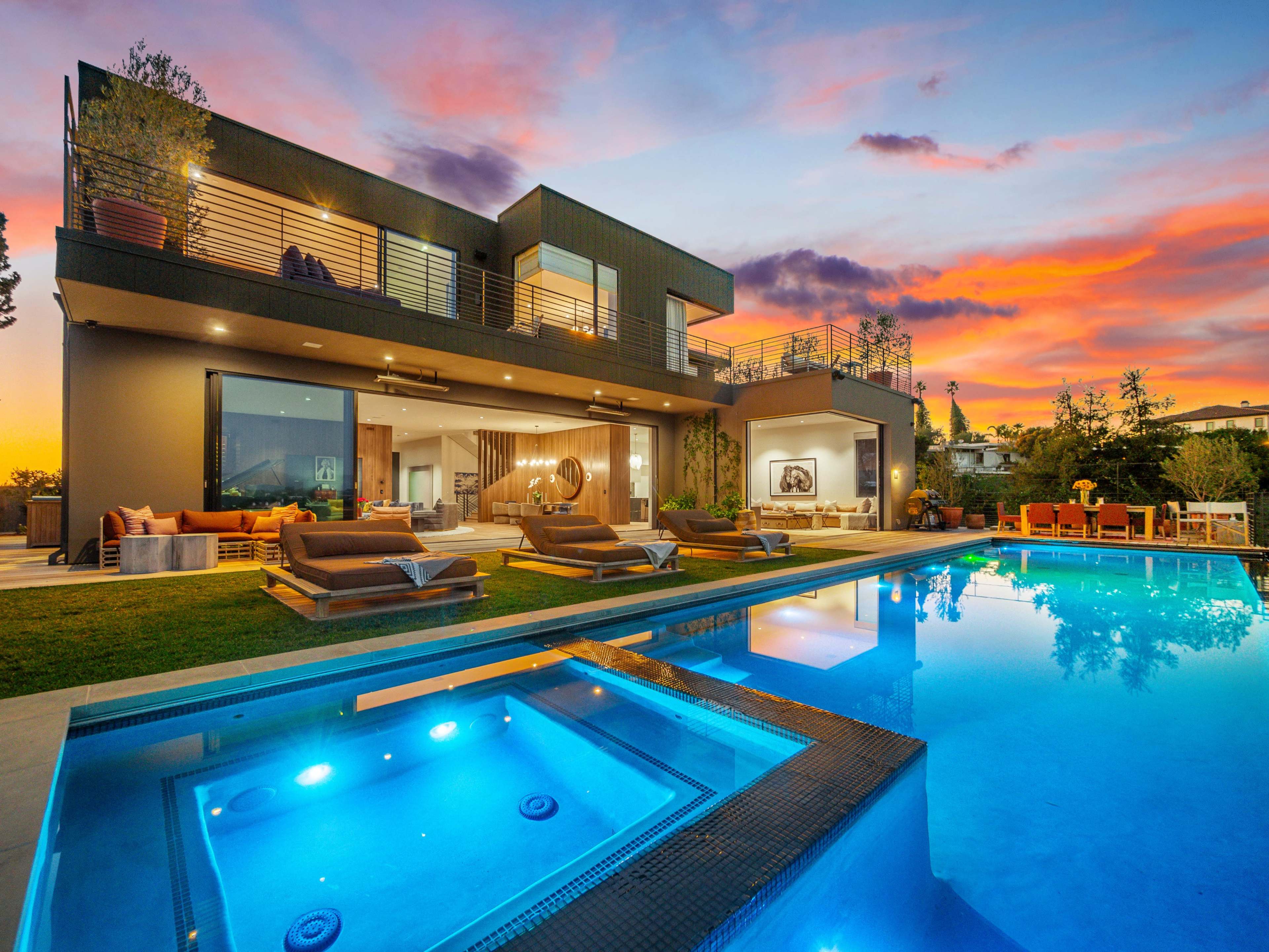 A modern two-story house with large glass windows and a pool in front reflects the colorful sunset sky.