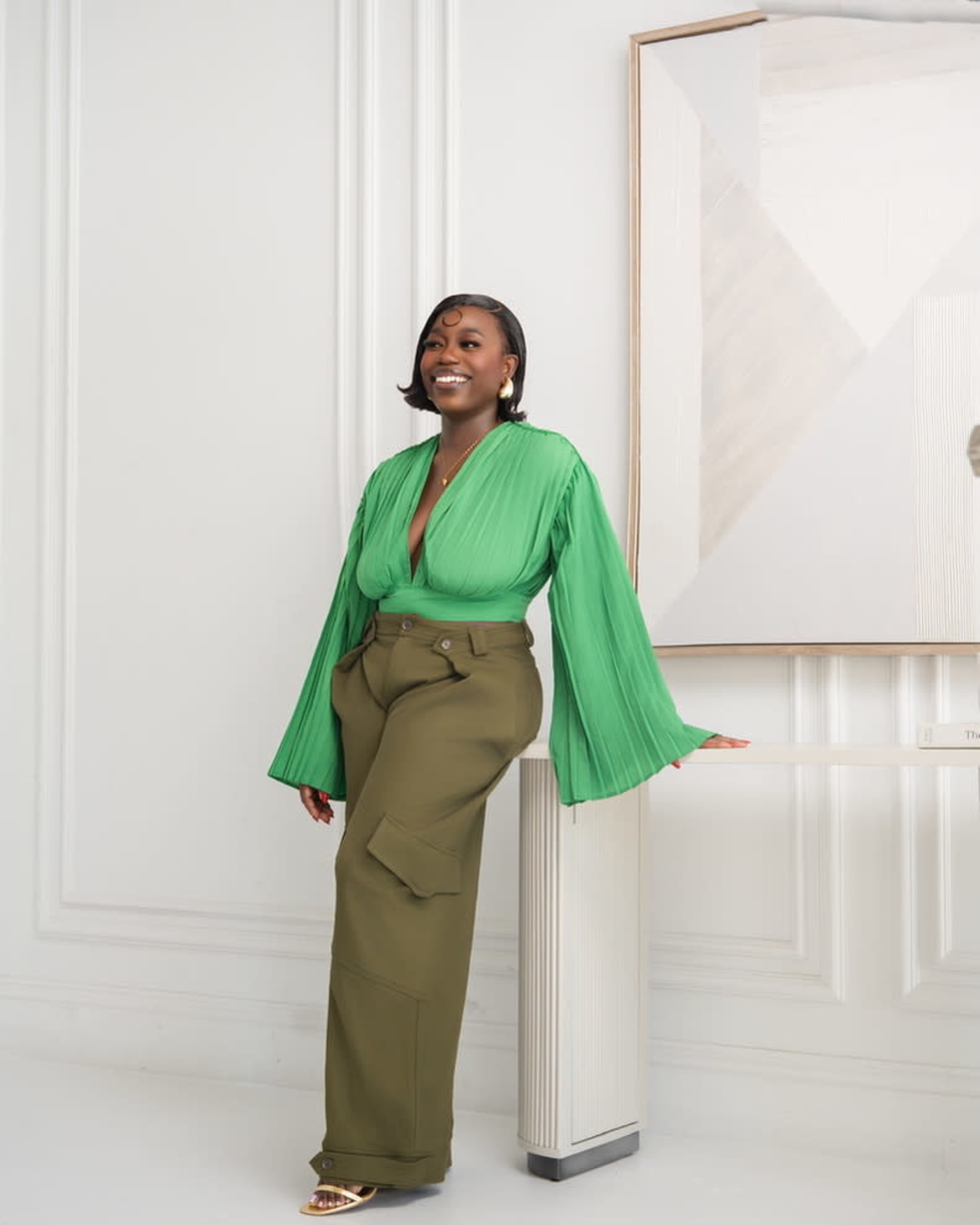 A woman in a green pleated blouse and olive green wide-leg pants stands next to a wall decoration in a well-lit interior space.