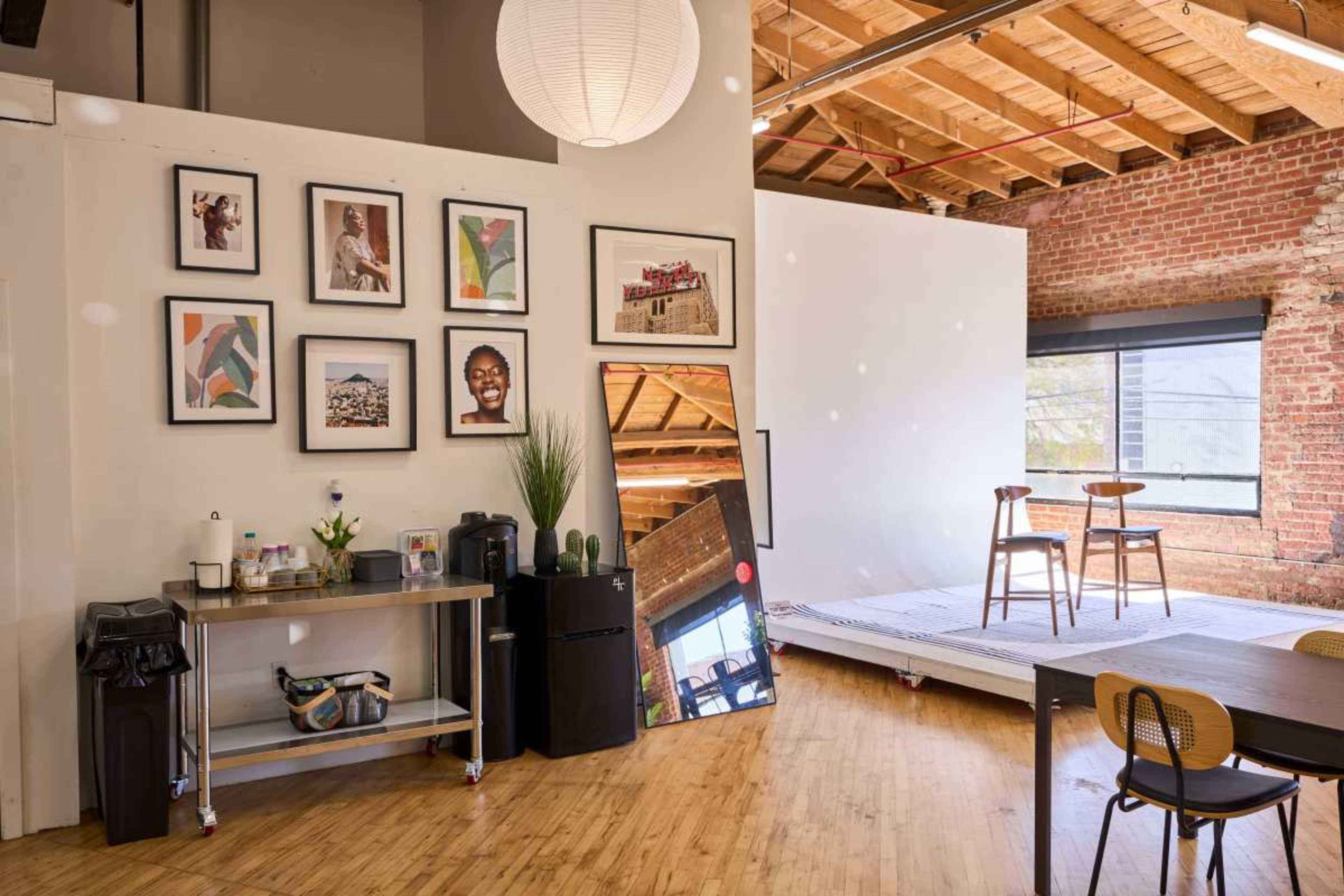 The image shows a spacious room featuring a collection of framed photos on one wall, a kitchenette area with appliances, and a small performance stage with two chairs in the background.