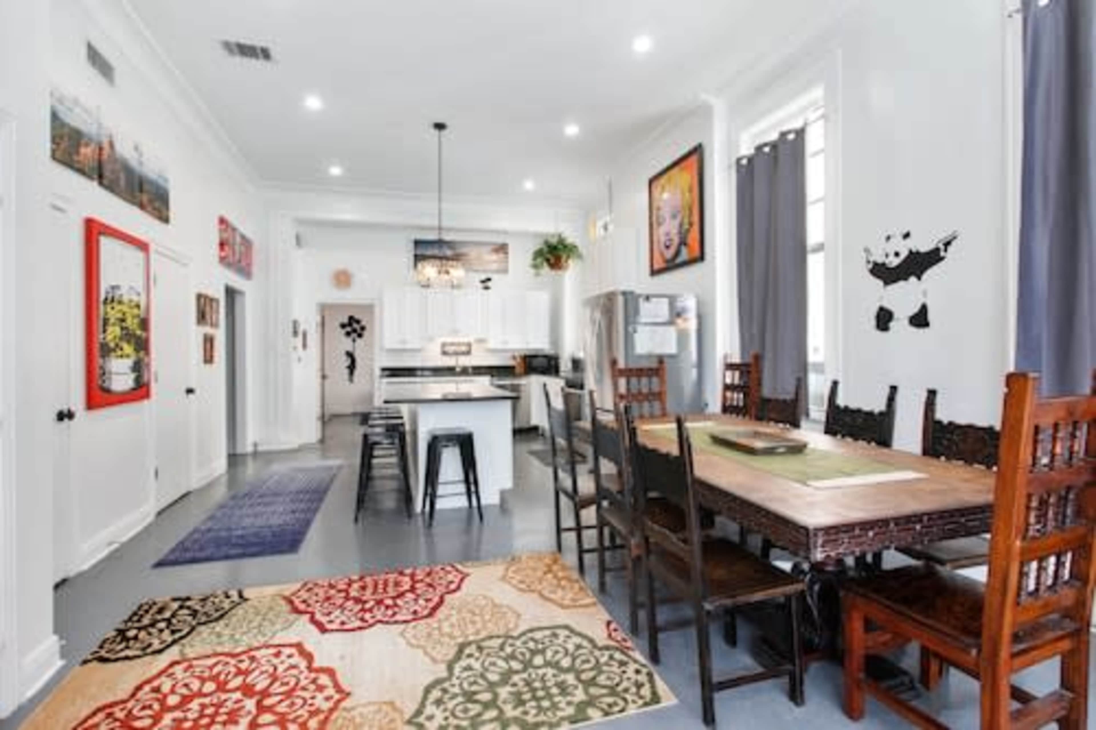 A spacious kitchen and dining area features a long wooden table surrounded by dark chairs, with colorful rugs and artwork on the walls.