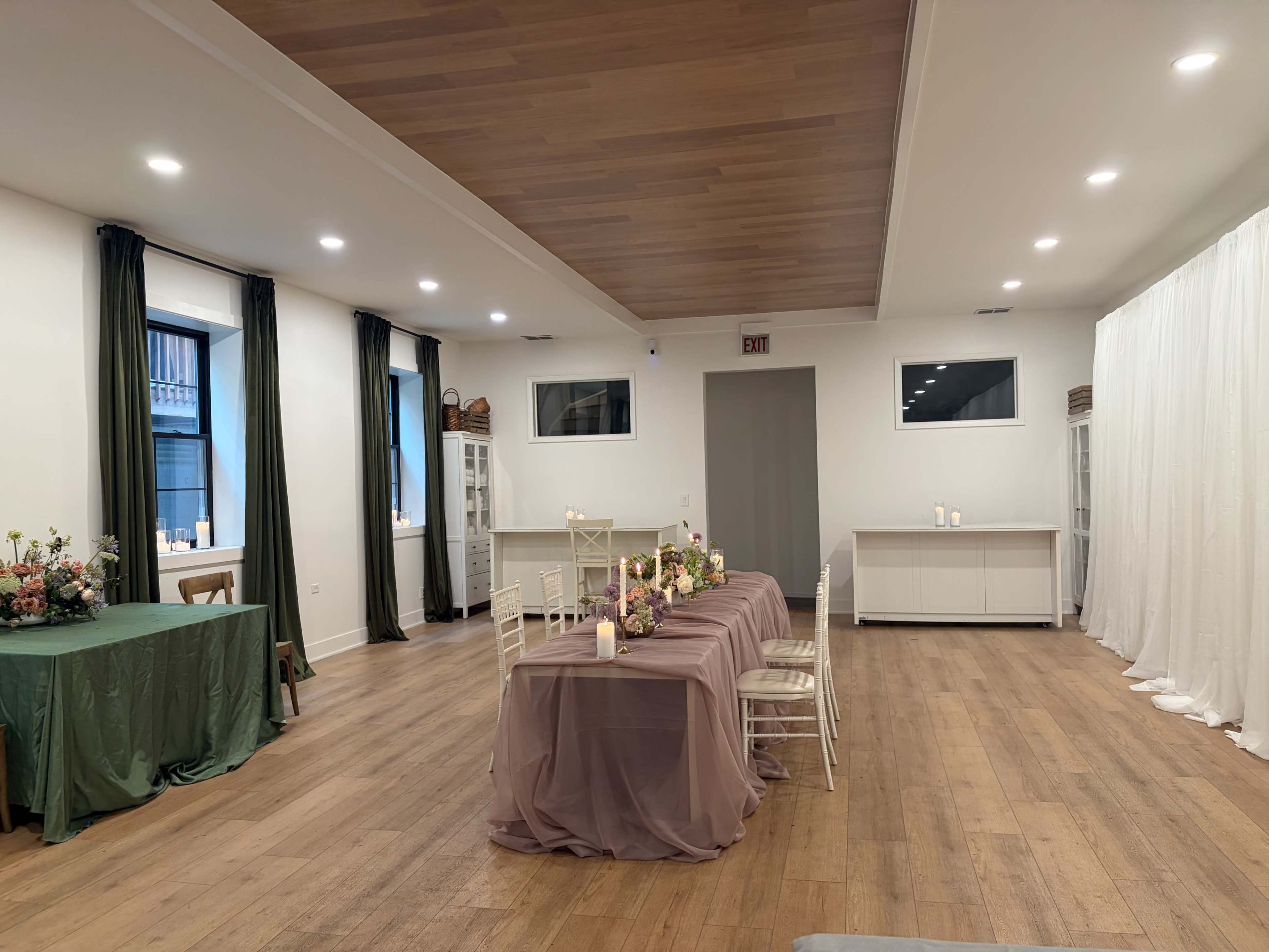 The image shows a spacious room with wooden flooring, featuring a long table set with a lavender tablecloth and chairs, flanked by two green tables and a white backdrop on one side.