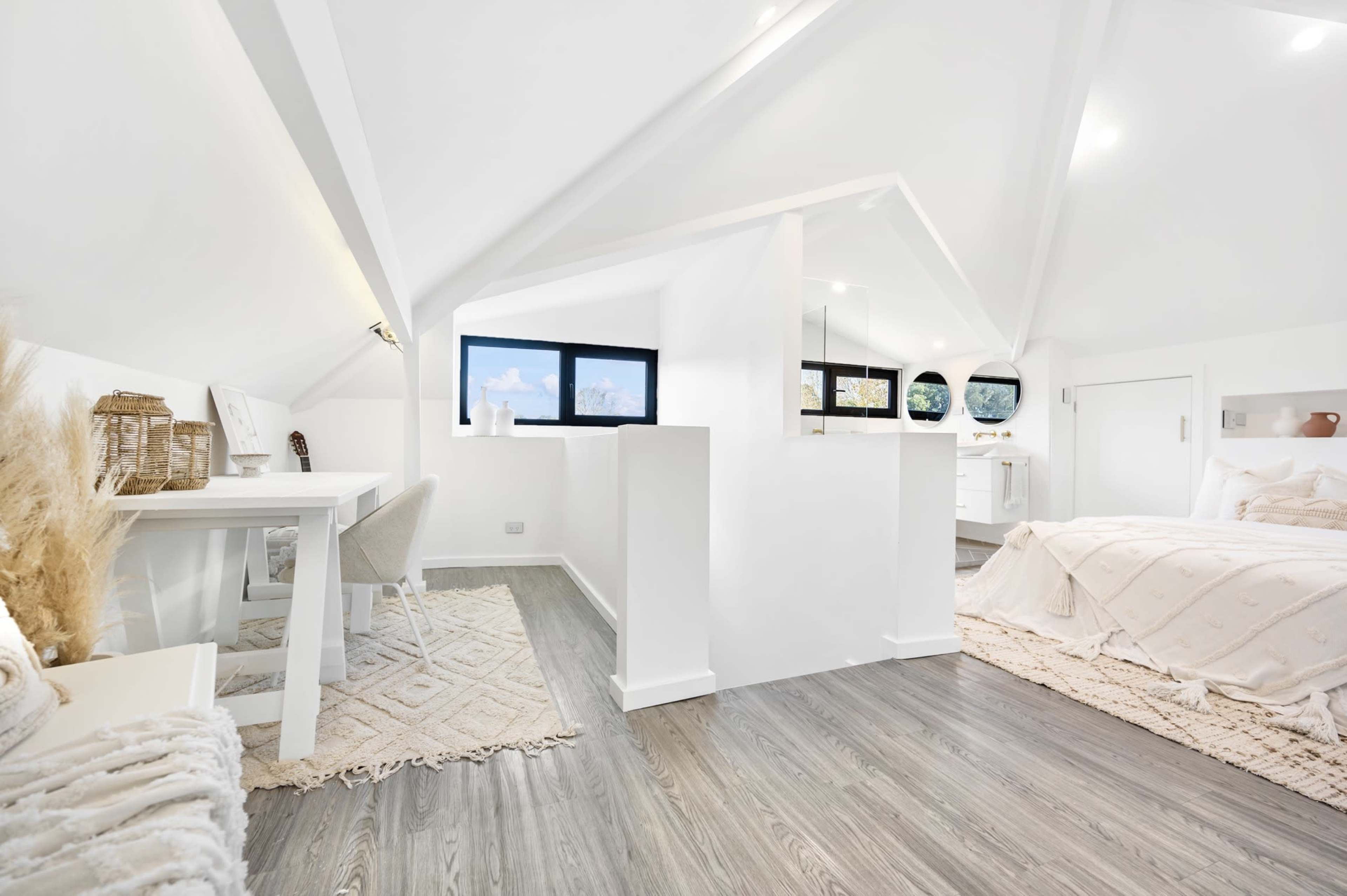 A bright, modern attic space featuring a desk area, a bed, and large windows that offer views of the outdoors.