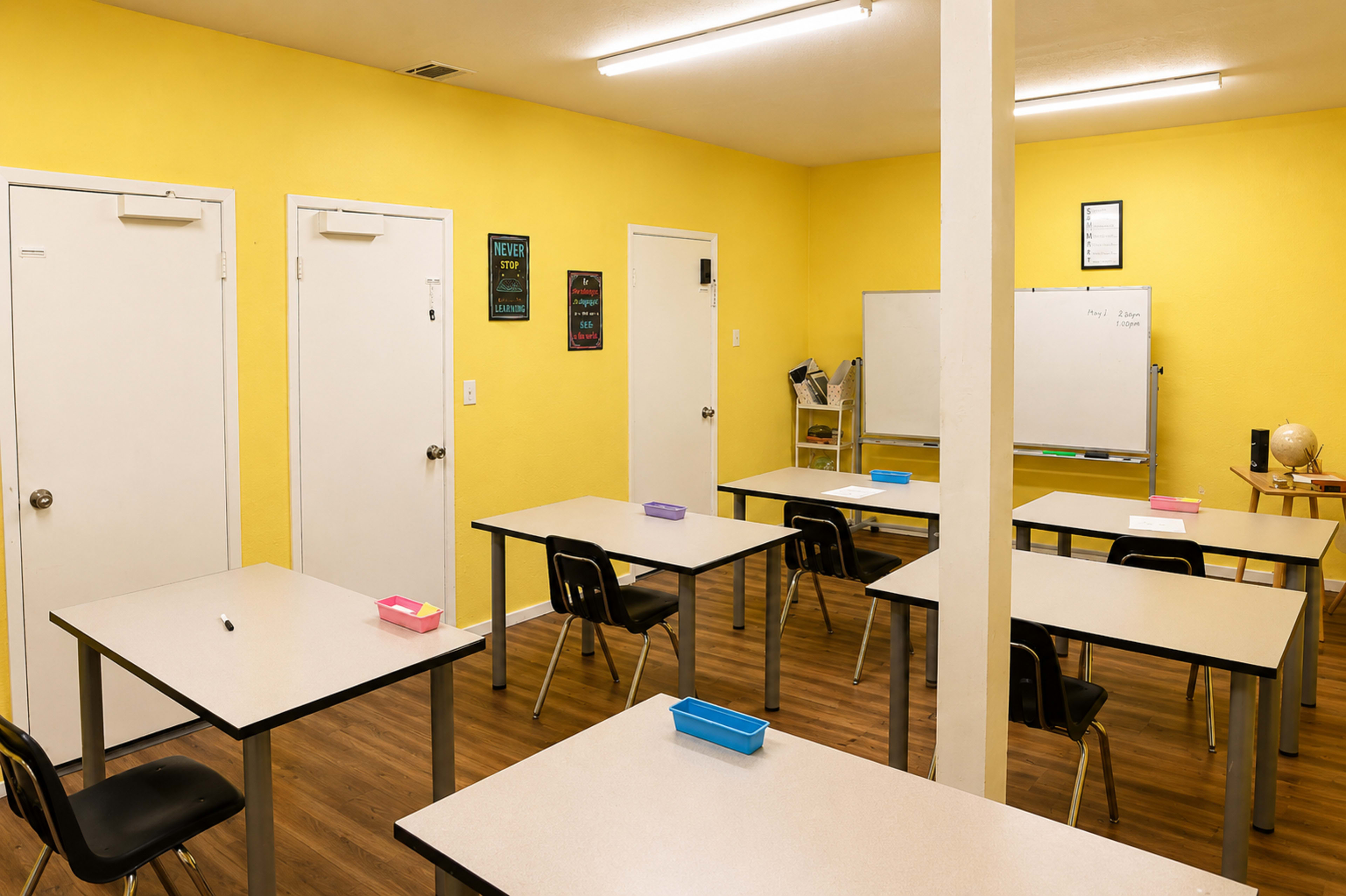 The image shows a classroom with yellow walls, featuring several tables and chairs arranged in a neat layout, along with a whiteboard and various doors.