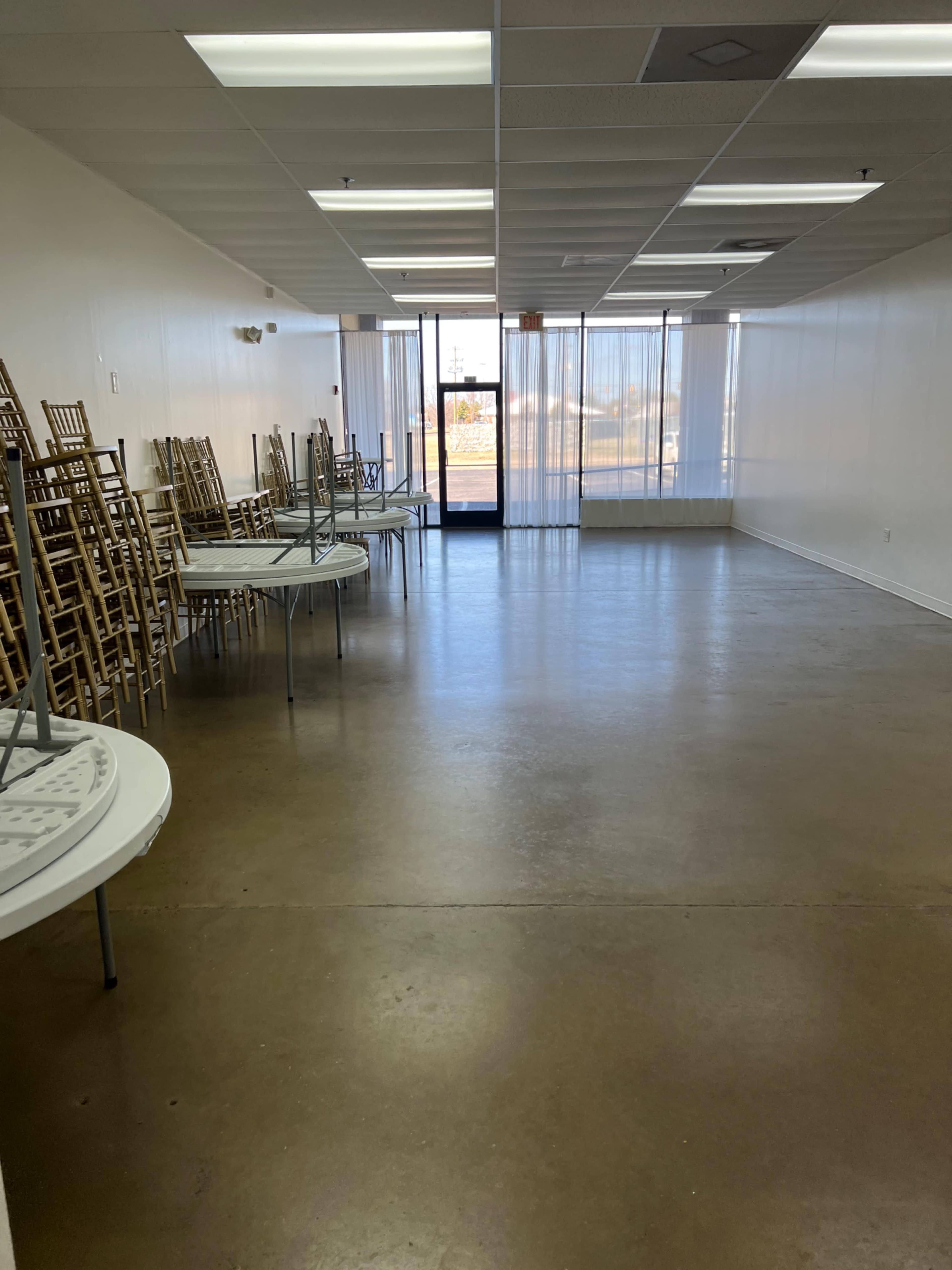 An empty room with a polished concrete floor, featuring several stacked chairs against the wall and tables turned upside down on circular bases.