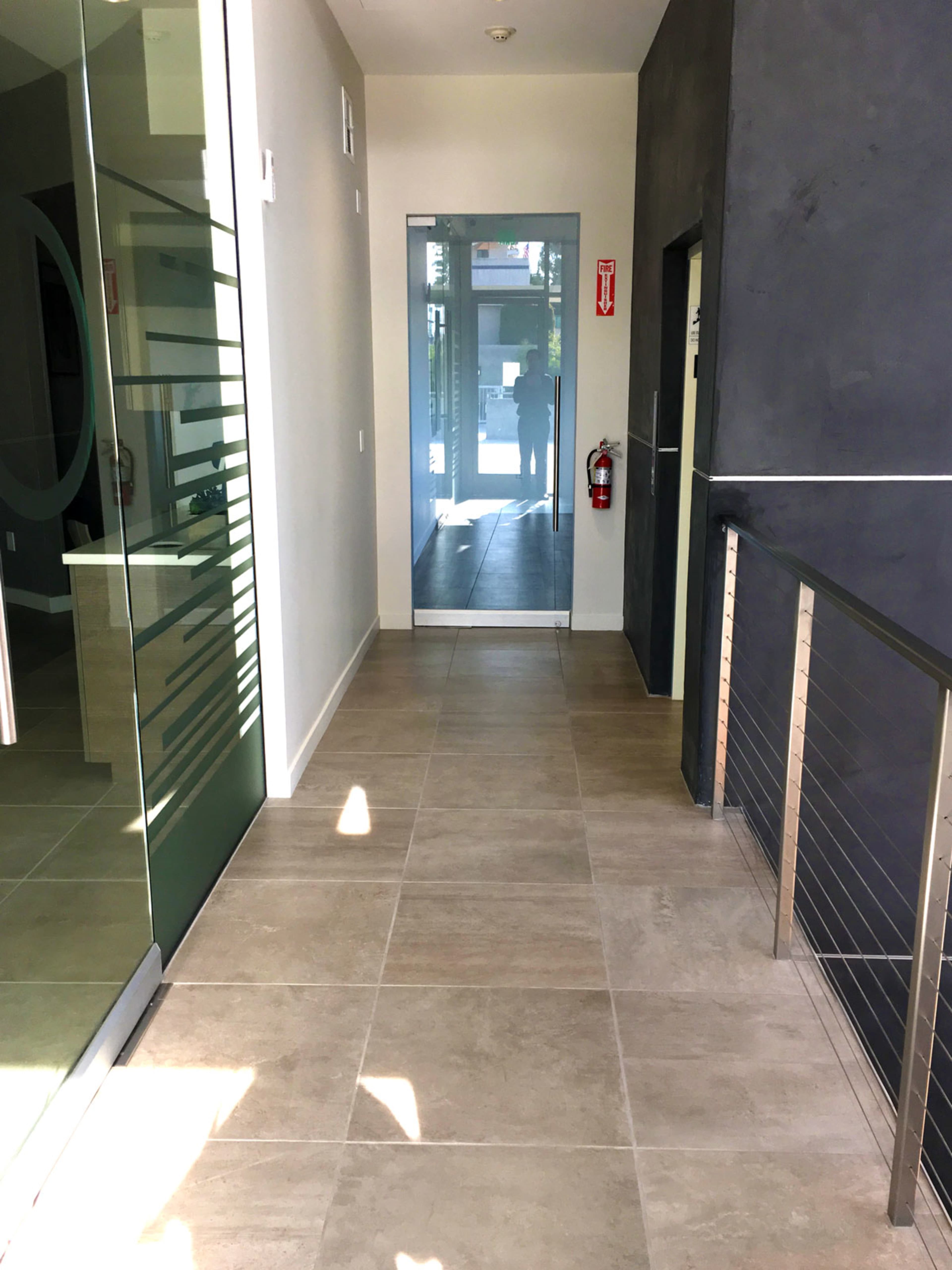 A corridor features tiled flooring, a glass partition on the left, and doors at both ends leading to outside views.