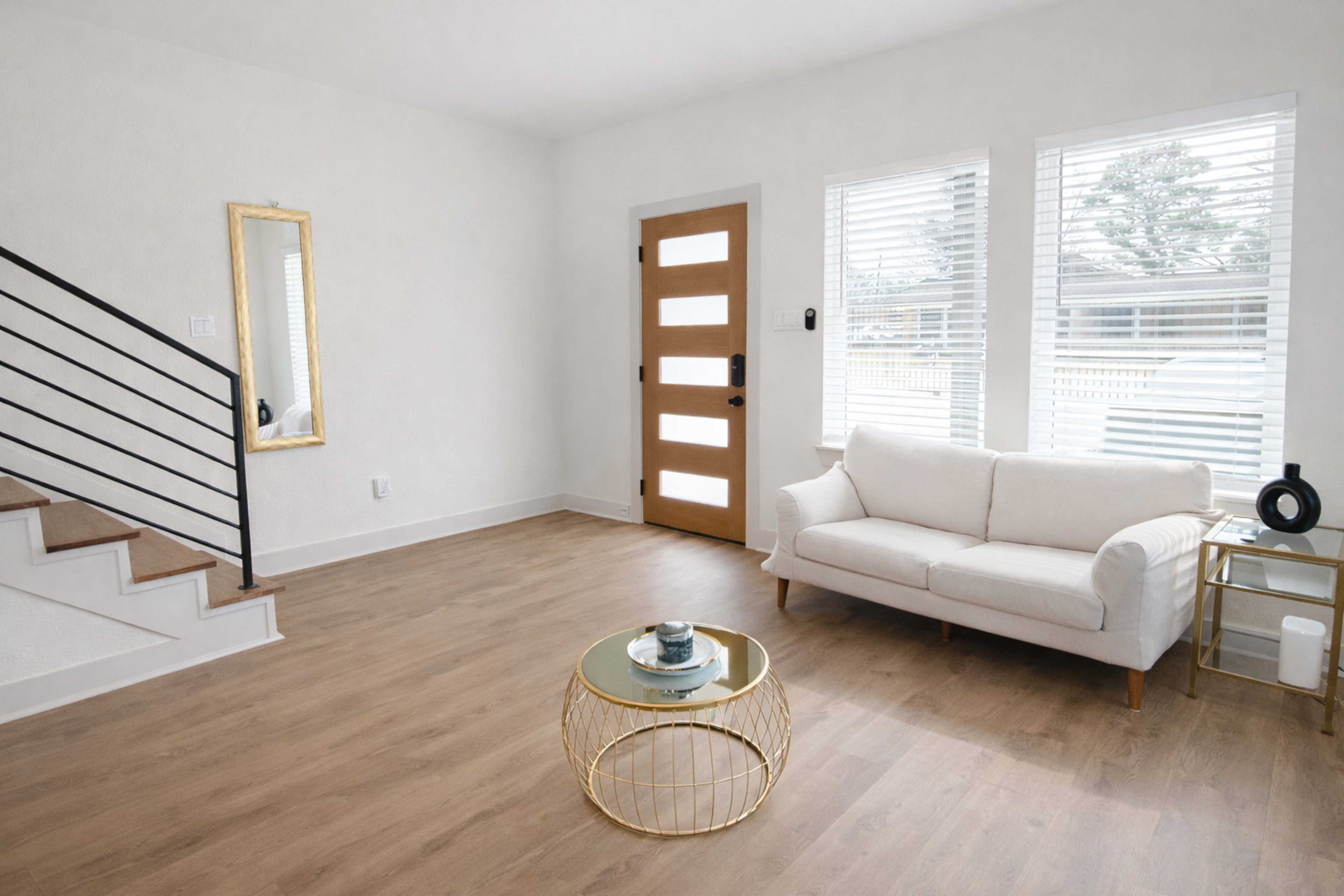 The scene features a bright, minimalist living room with a white couch, a round coffee table, a mirror, and a modern front door with rectangular windows.