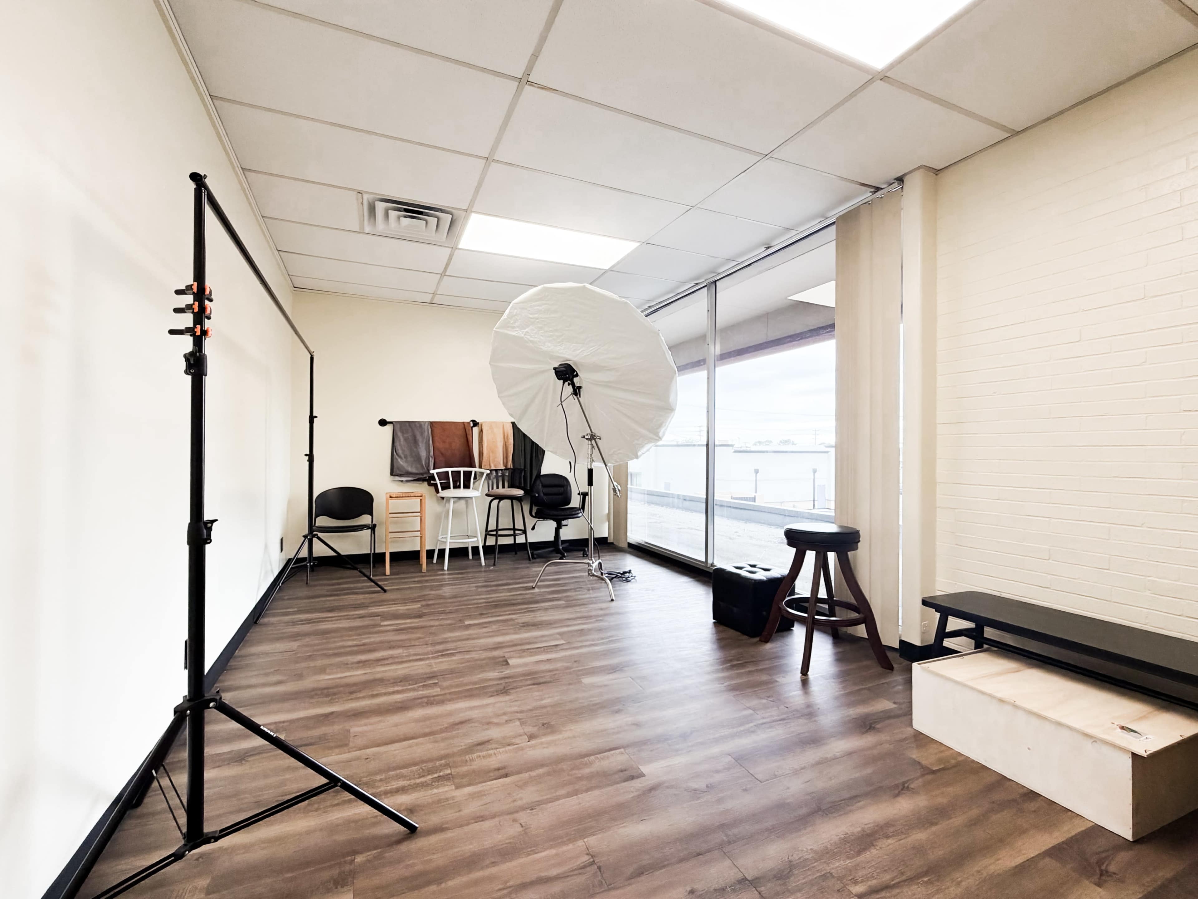 The image shows a spacious photography studio with a large softbox, a backdrop, and a few chairs arranged in the corner.