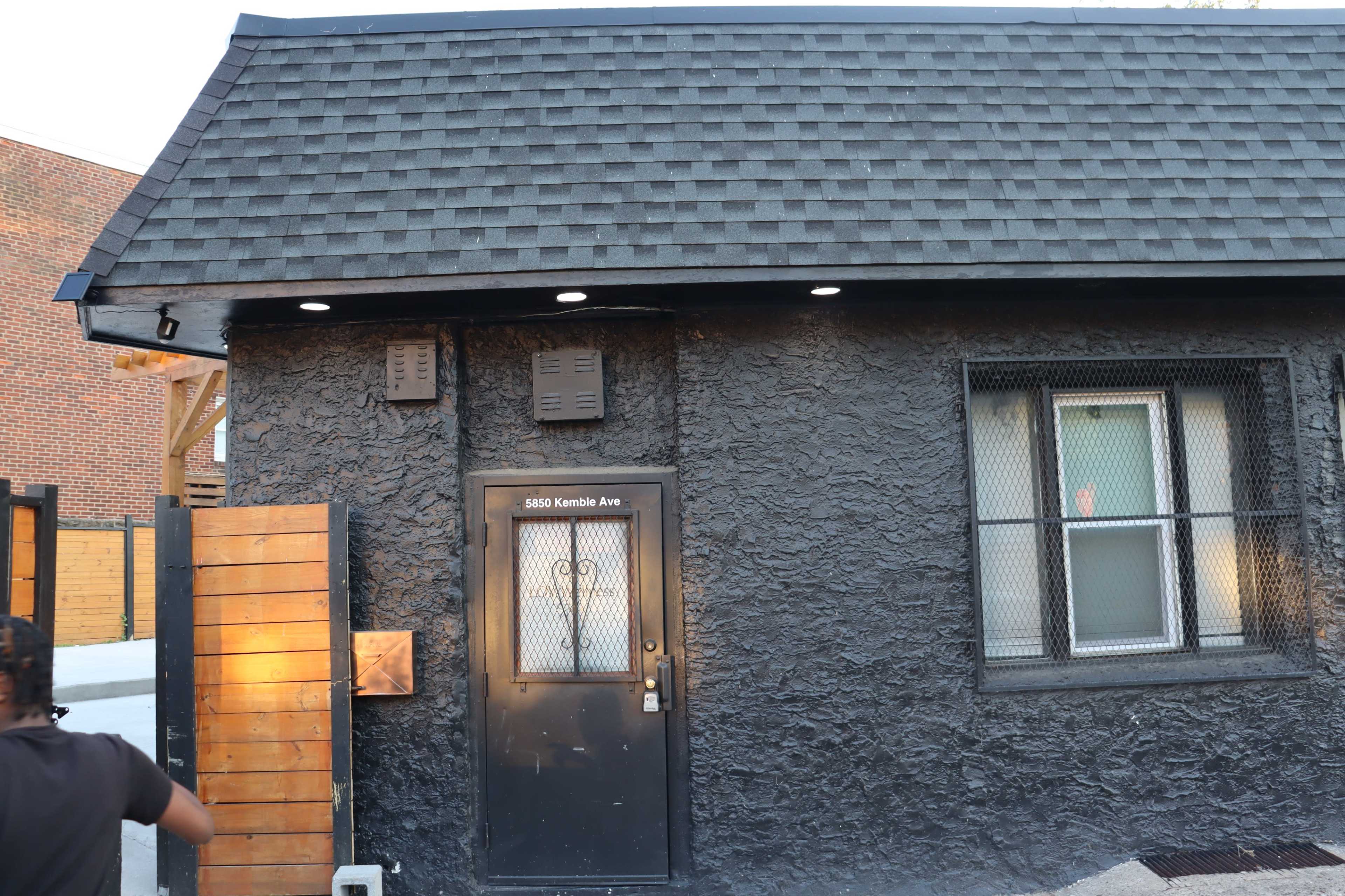 The image shows a black textured building with a sloped roof and a door featuring a metal grate.