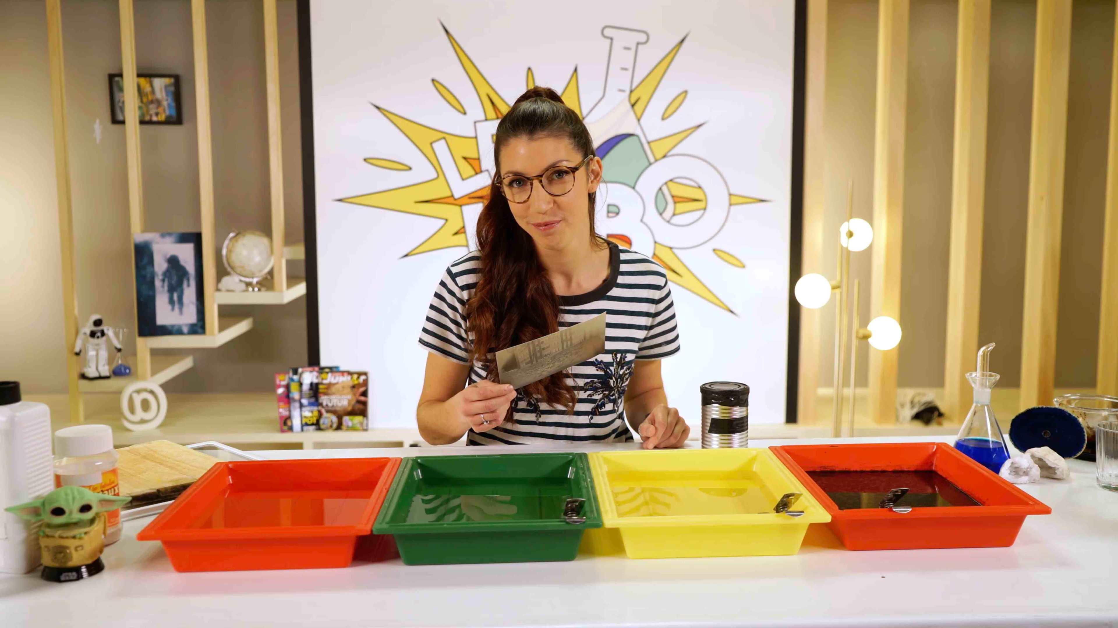 A woman wearing glasses holds a knife while sitting at a table with four colored trays filled with liquids.