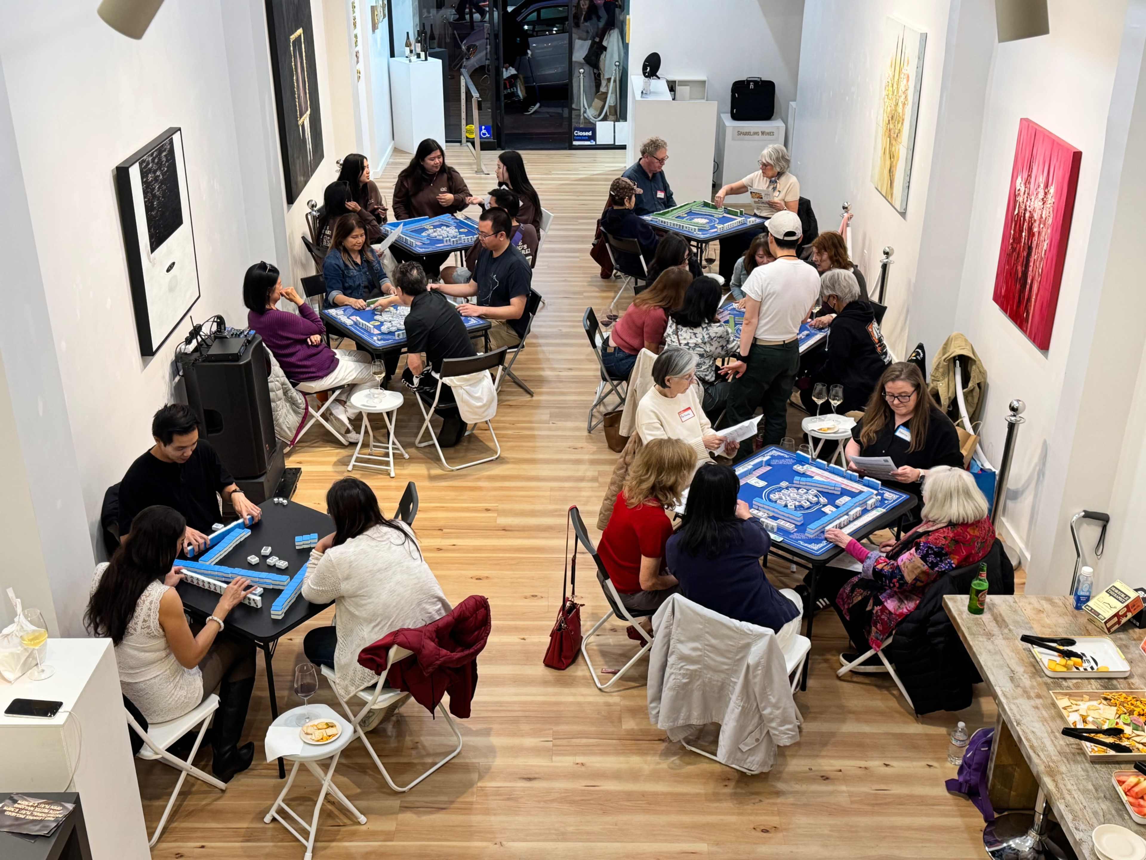 The image shows a group of people seated at tables in a spacious room, engaged in playing board games.