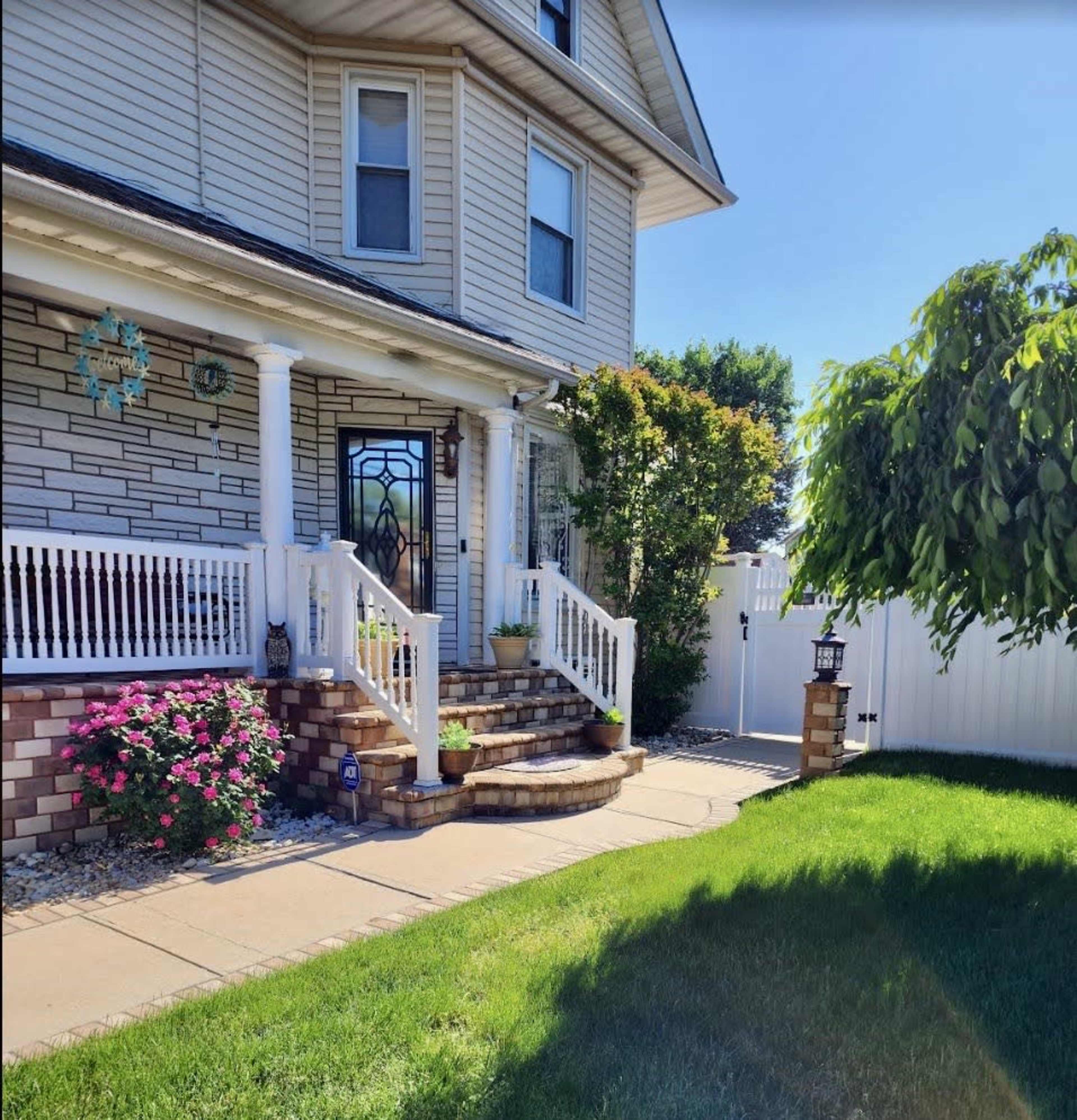 A two-story house with a covered front porch, surrounded by greenery and colorful flowers along the walkway.