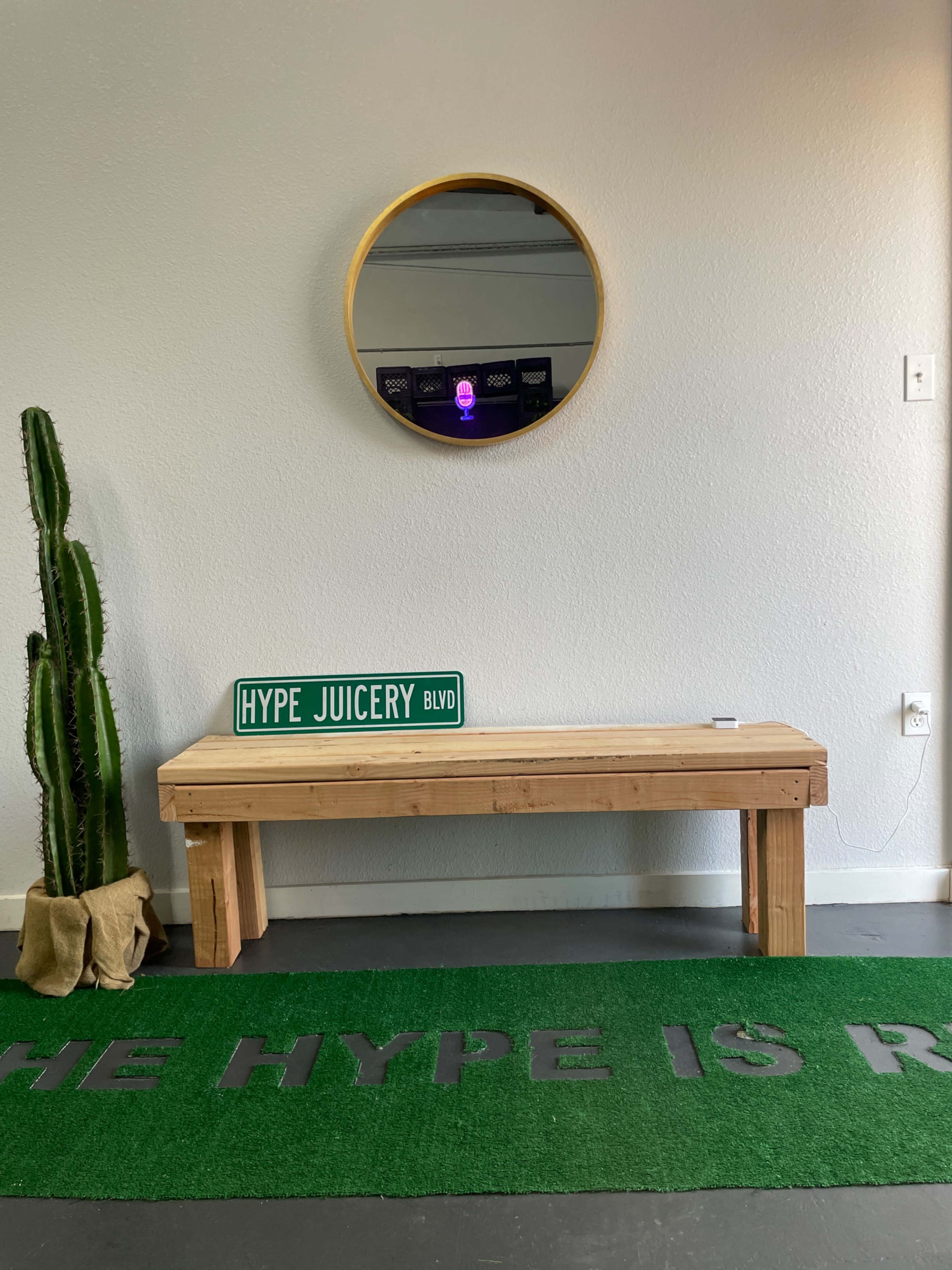 A wooden bench with a sign reading "HYPE JUICERY BLVD," a round mirror on the wall, and a large green rug with the text "THE HYPE IS REAL" in a modern interior space.