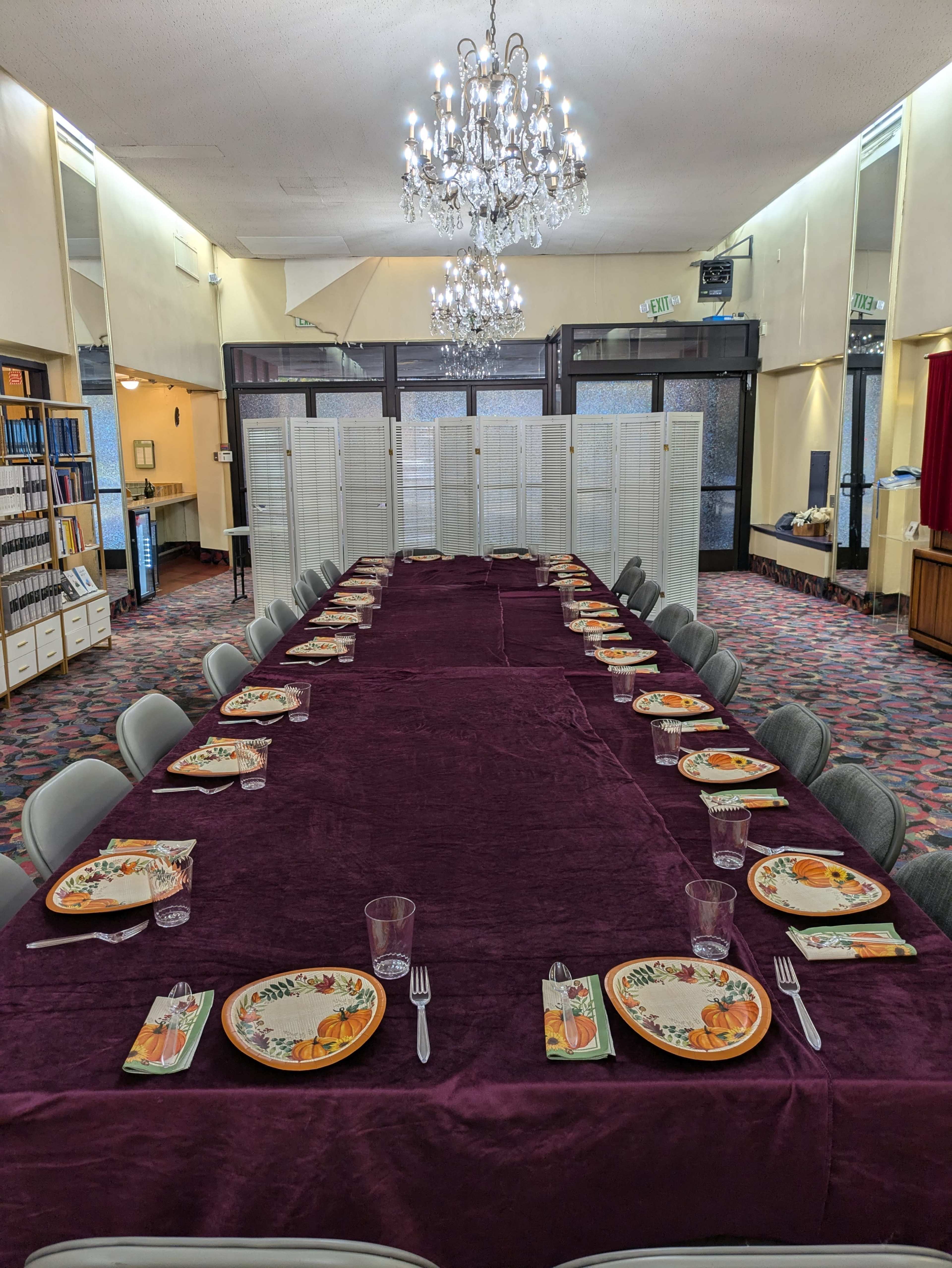 A long, rectangular dining table is set with plates and utensils, draped in a purple tablecloth, beneath a chandelier in a spacious room divided by folding screens.