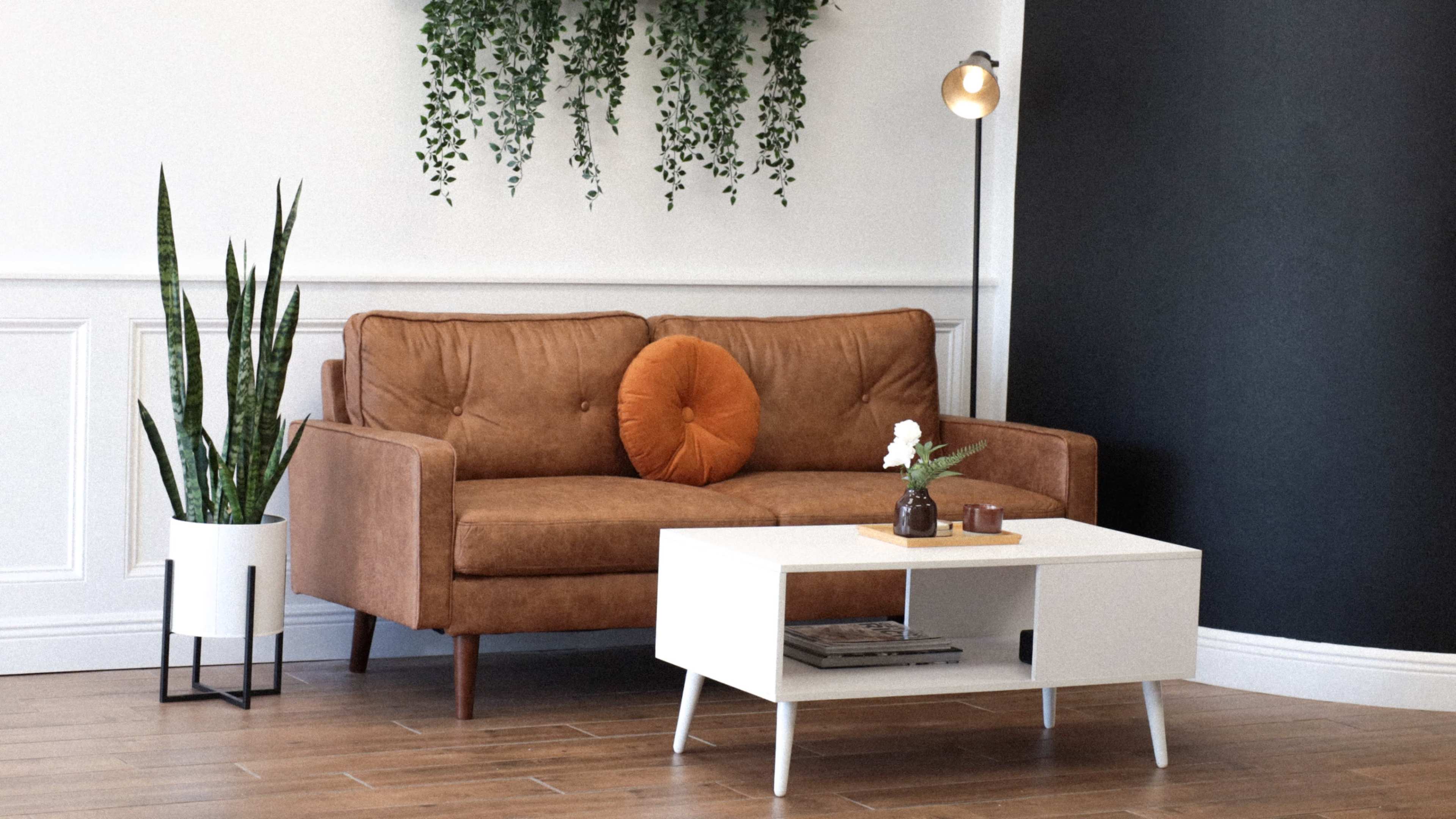 A brown fabric sofa with an orange pillow sits opposite a white coffee table, surrounded by indoor plants and a lamp, in a well-lit room with a black accent wall.