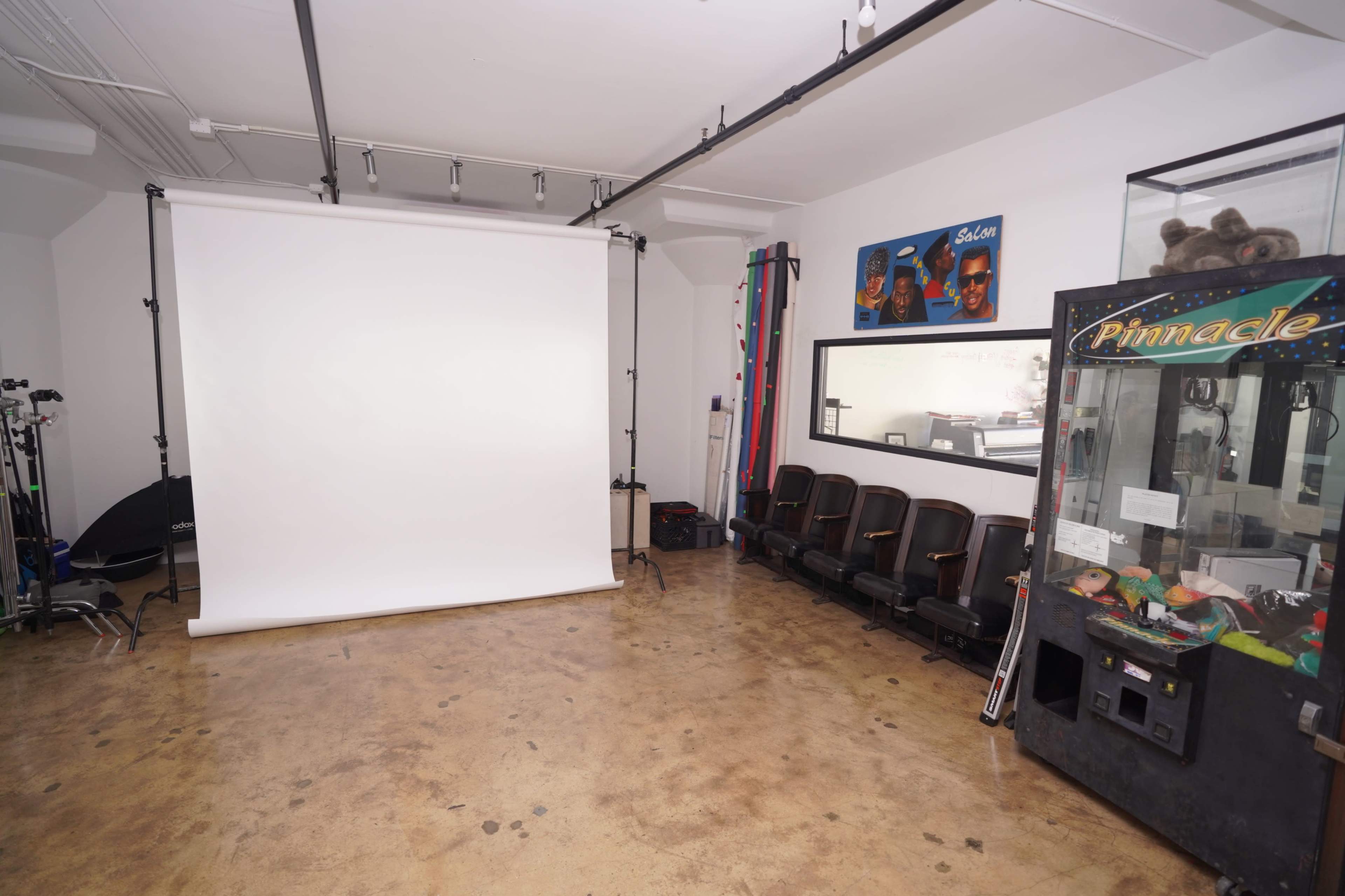 The image shows a photography studio featuring a large white backdrop, portable lighting equipment, and a claw machine against one wall.