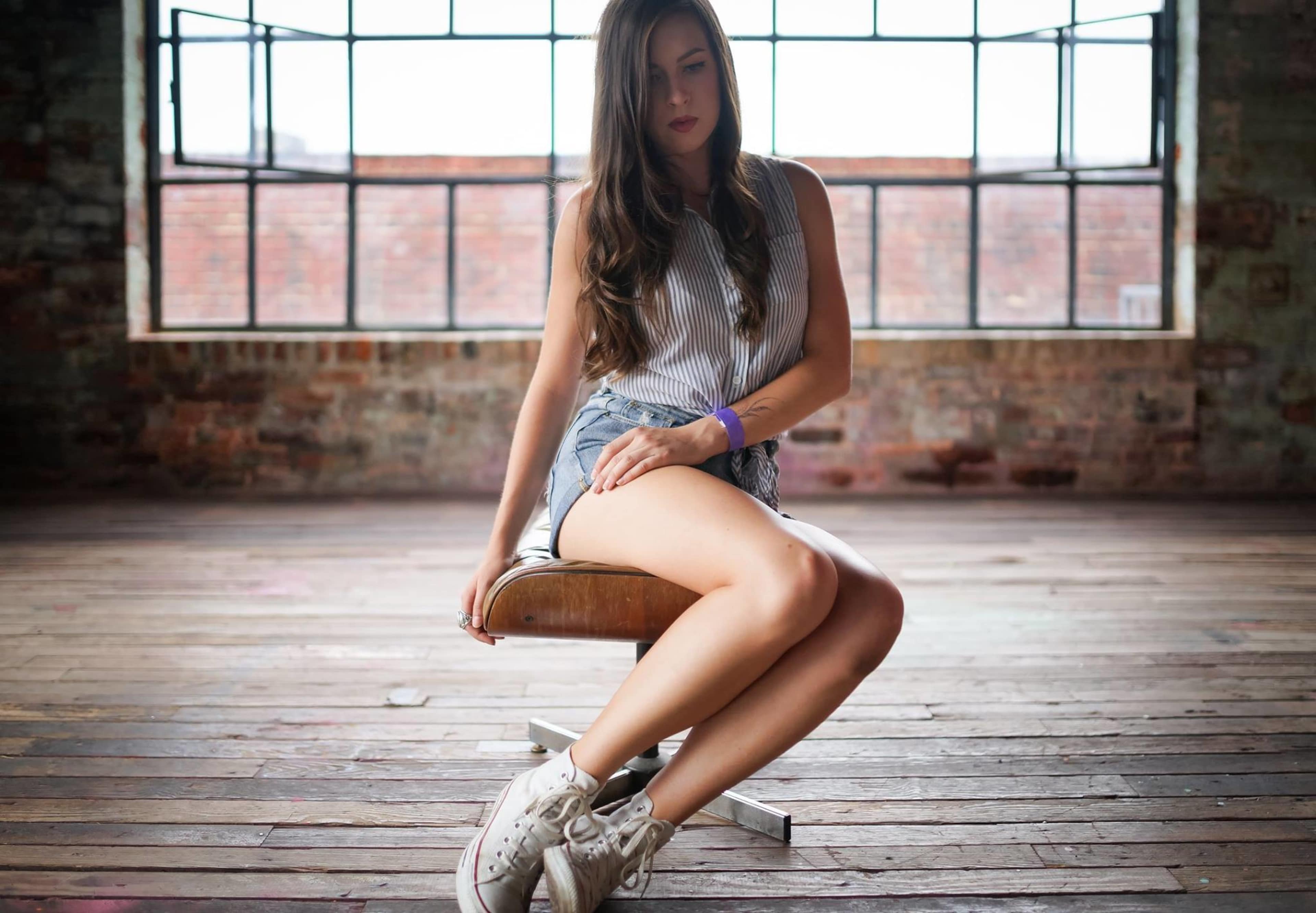 A woman sits on a wooden stool in a spacious, industrial-style room with large windows.