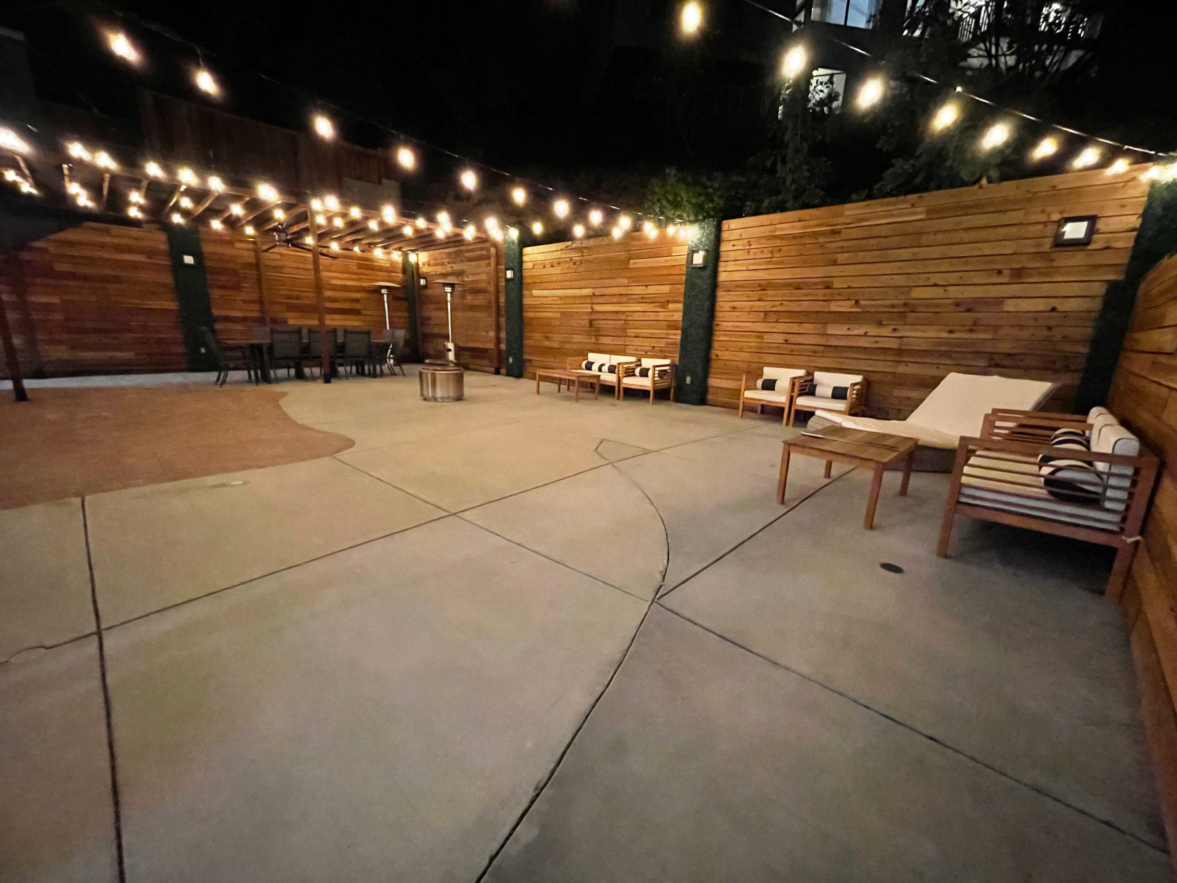 A dimly lit outdoor seating area with wooden walls, string lights overhead, and furniture arranged around a paved surface.