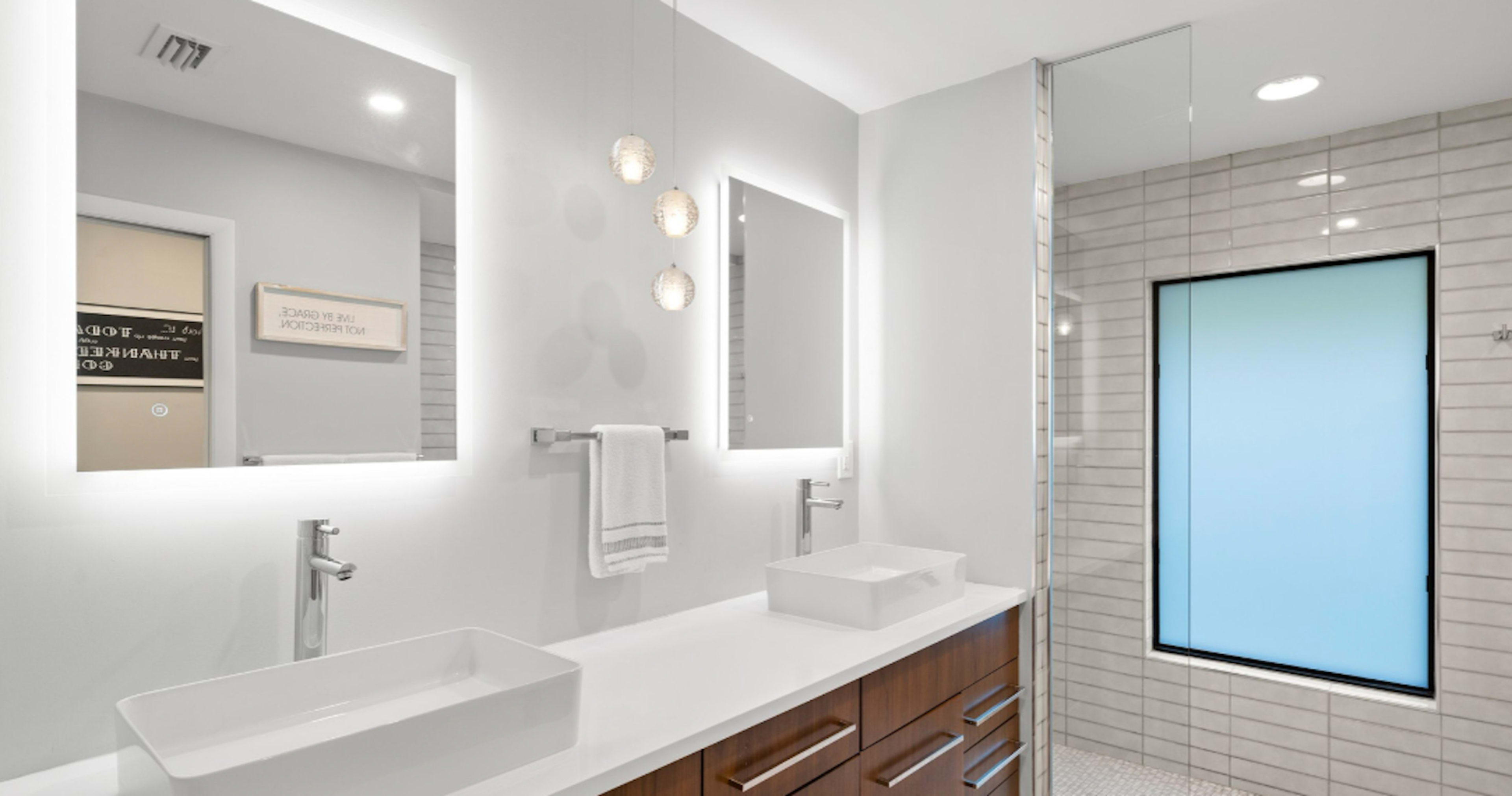 A modern bathroom features dual rectangular mirrors with integrated lighting above a white countertop and wooden drawers, alongside a walk-in shower with a frosted glass panel.