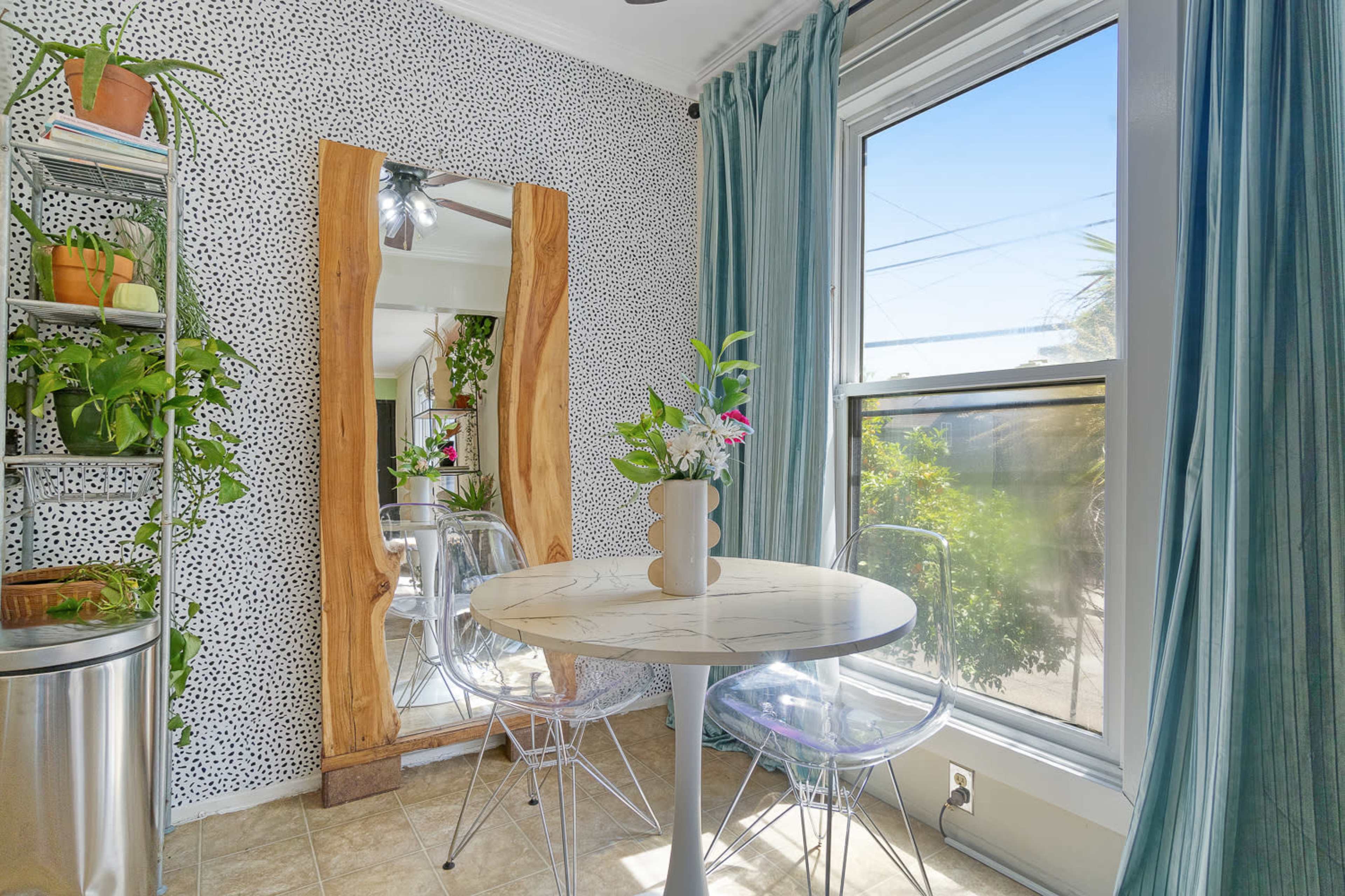 A small dining area features a round marble-top table surrounded by clear plastic chairs, with green plants and a large wooden mirror against a speckled wall.