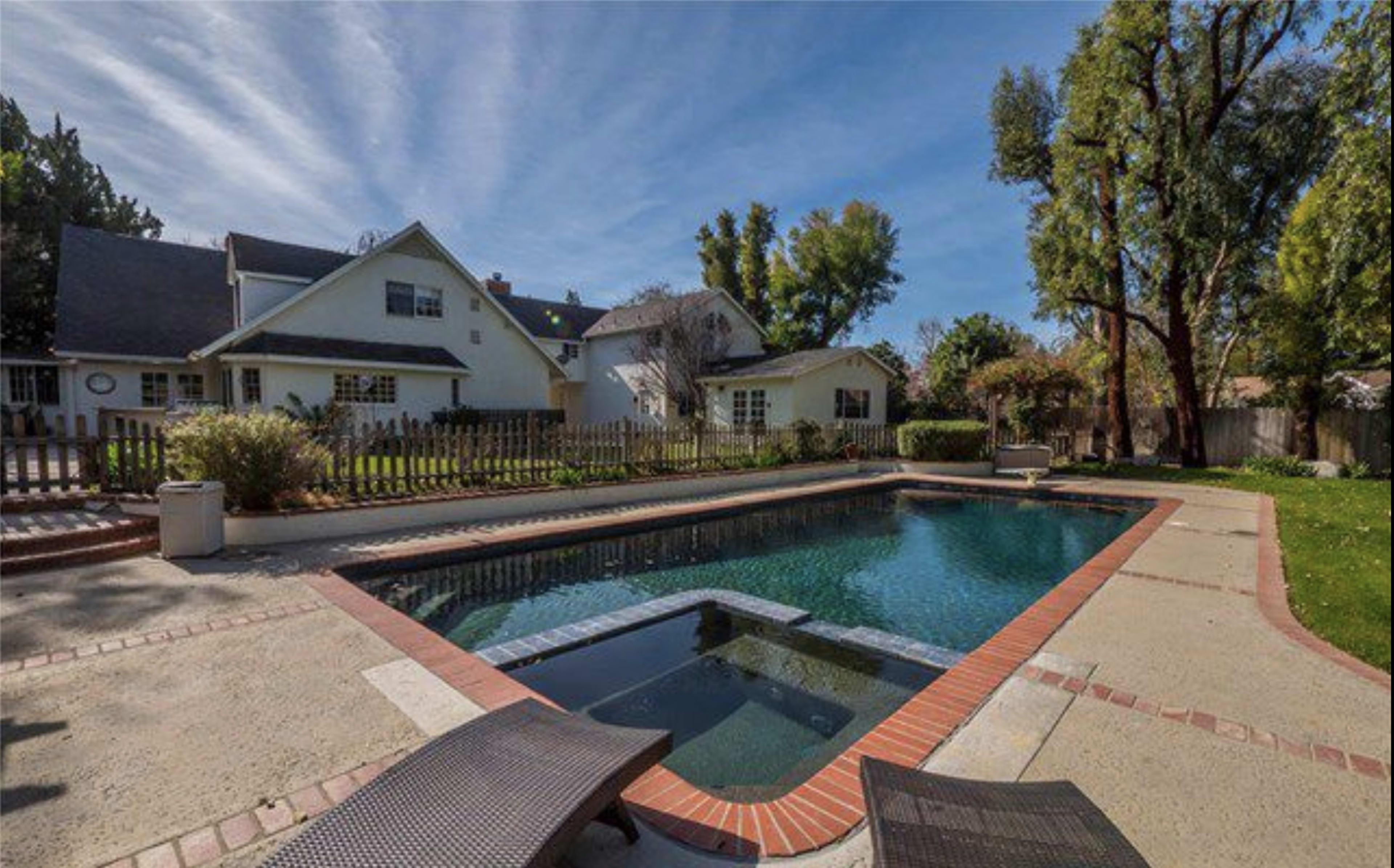 A clear swimming pool is surrounded by a concrete deck and grass, with a house and trees visible in the background.