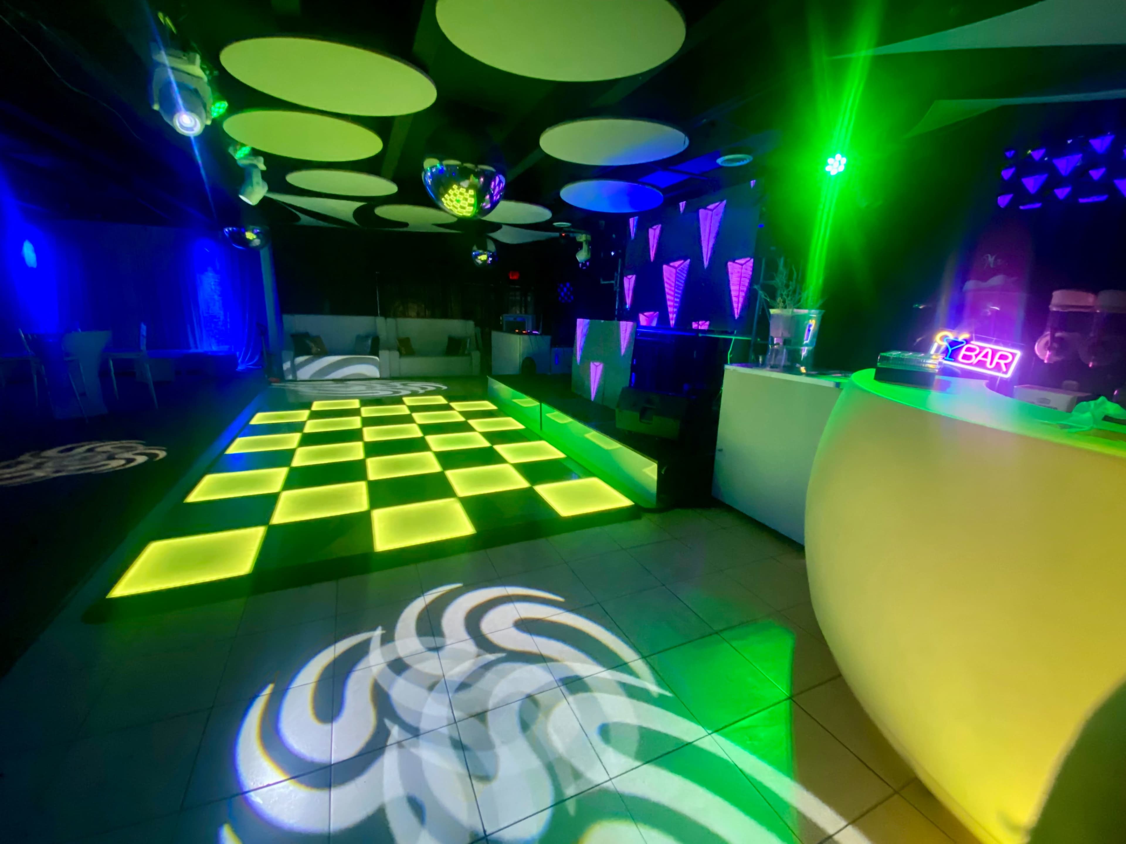 A nightclub interior featuring a checkerboard dance floor illuminated by green and blue lighting, with a bar area in the background.