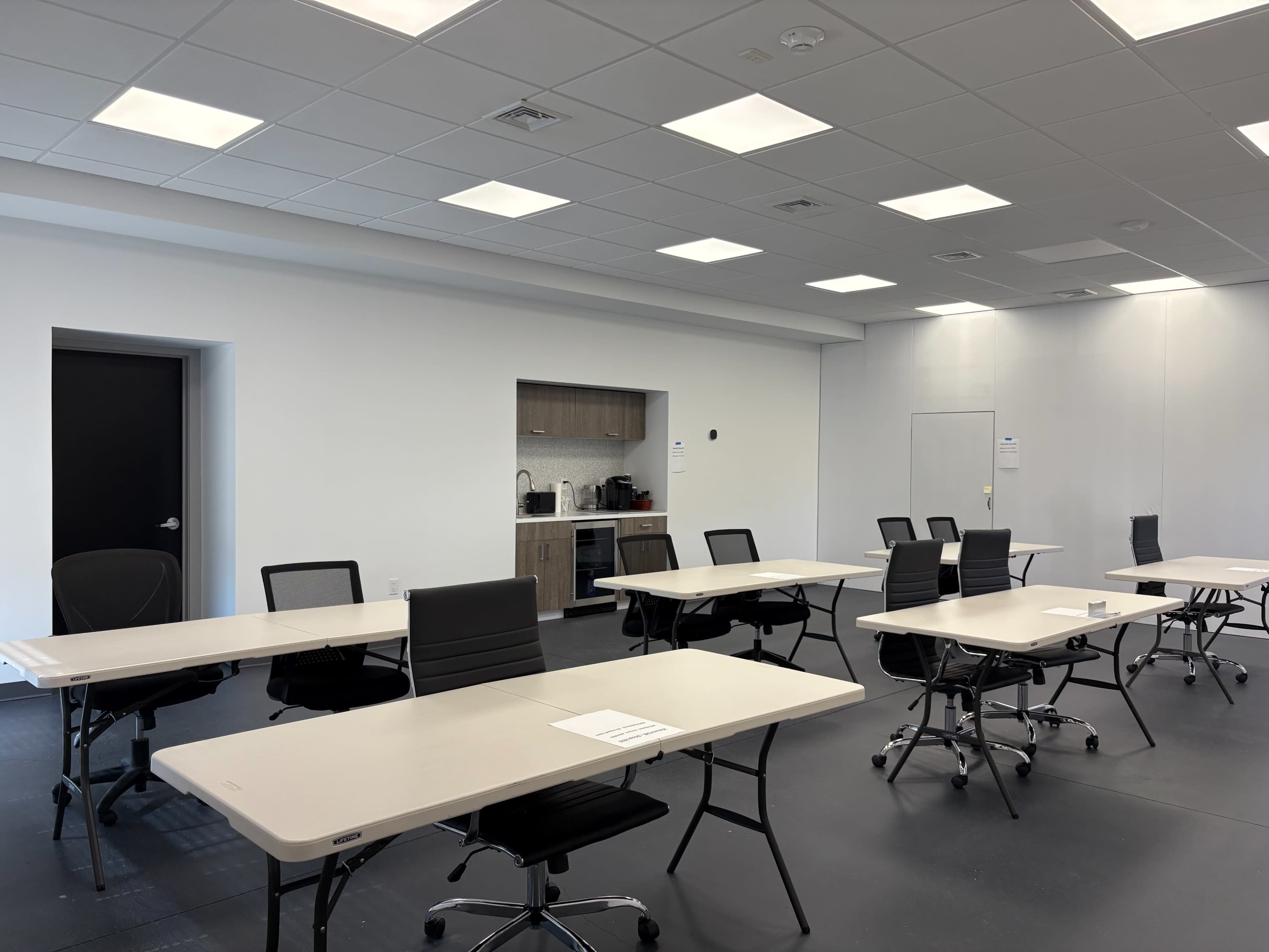 The image shows a large, empty room with several tables and chairs arranged for a meeting, along with a kitchenette area in the background.