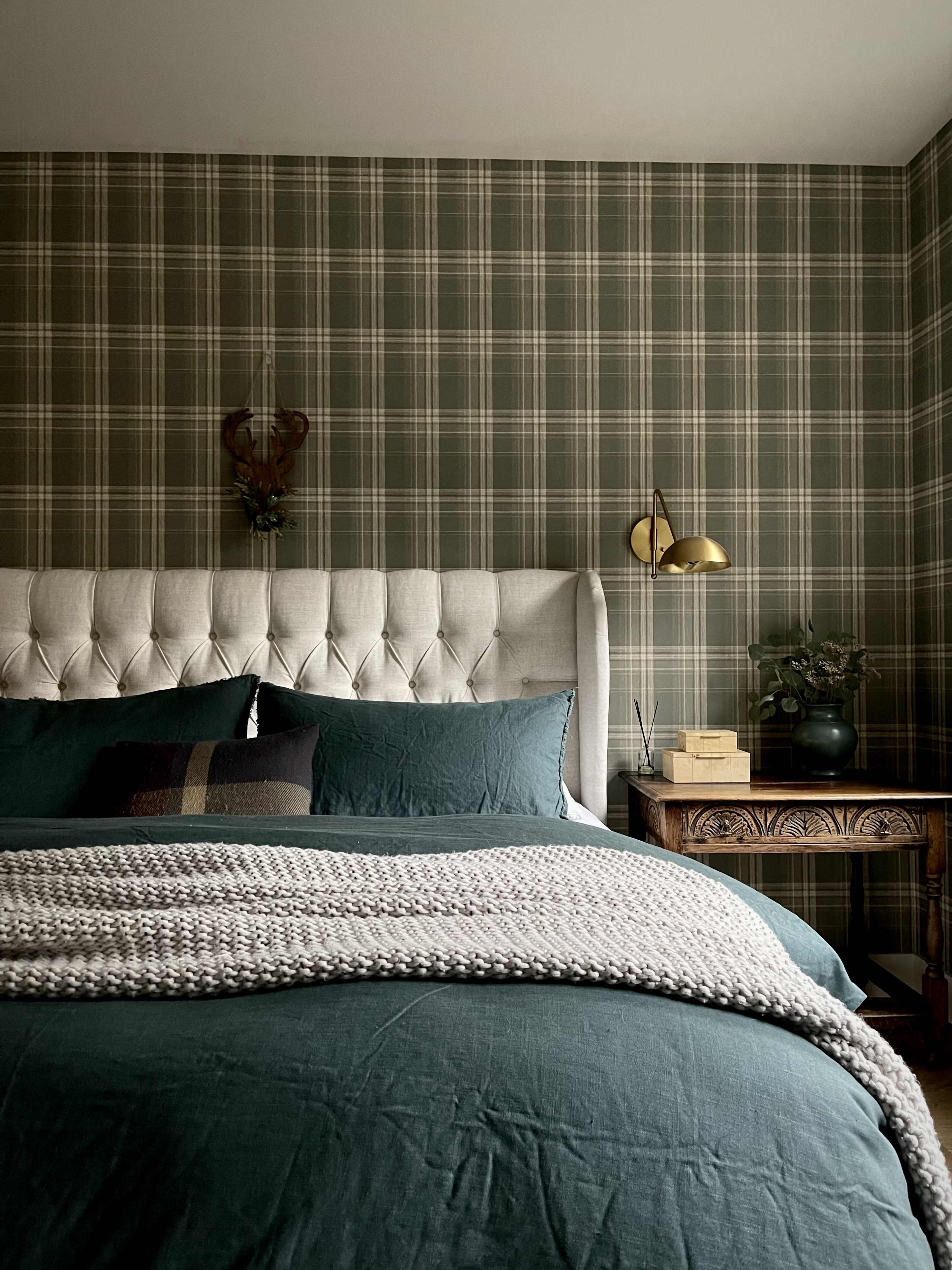 A neatly made bed with a tufted headboard and cozy blankets is positioned against a plaid wallpapered wall, accompanied by a side table and decorative elements.