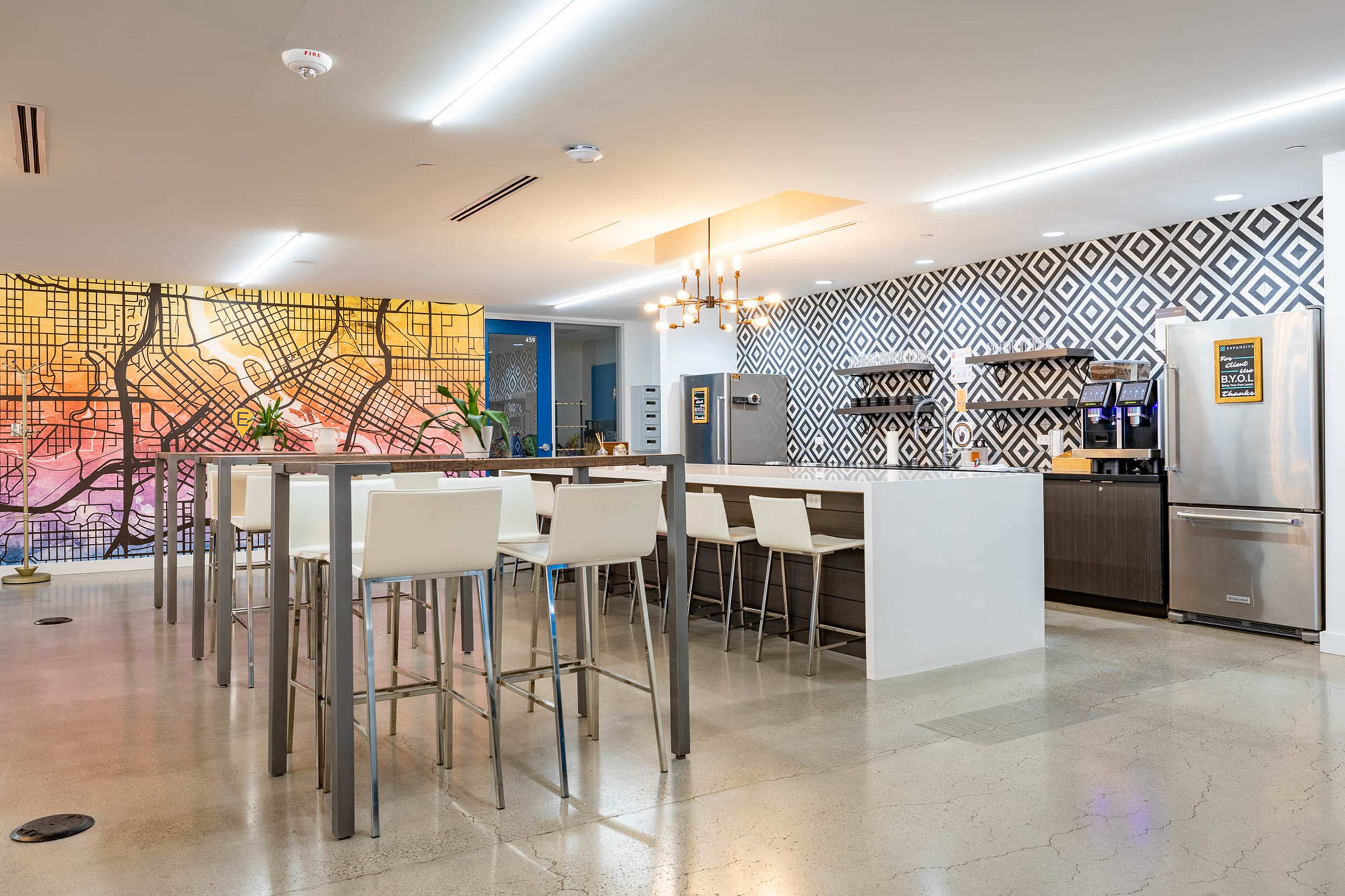 The image shows a modern kitchen area with a large communal table, high chairs, and a decorative wall featuring a colorful map design.