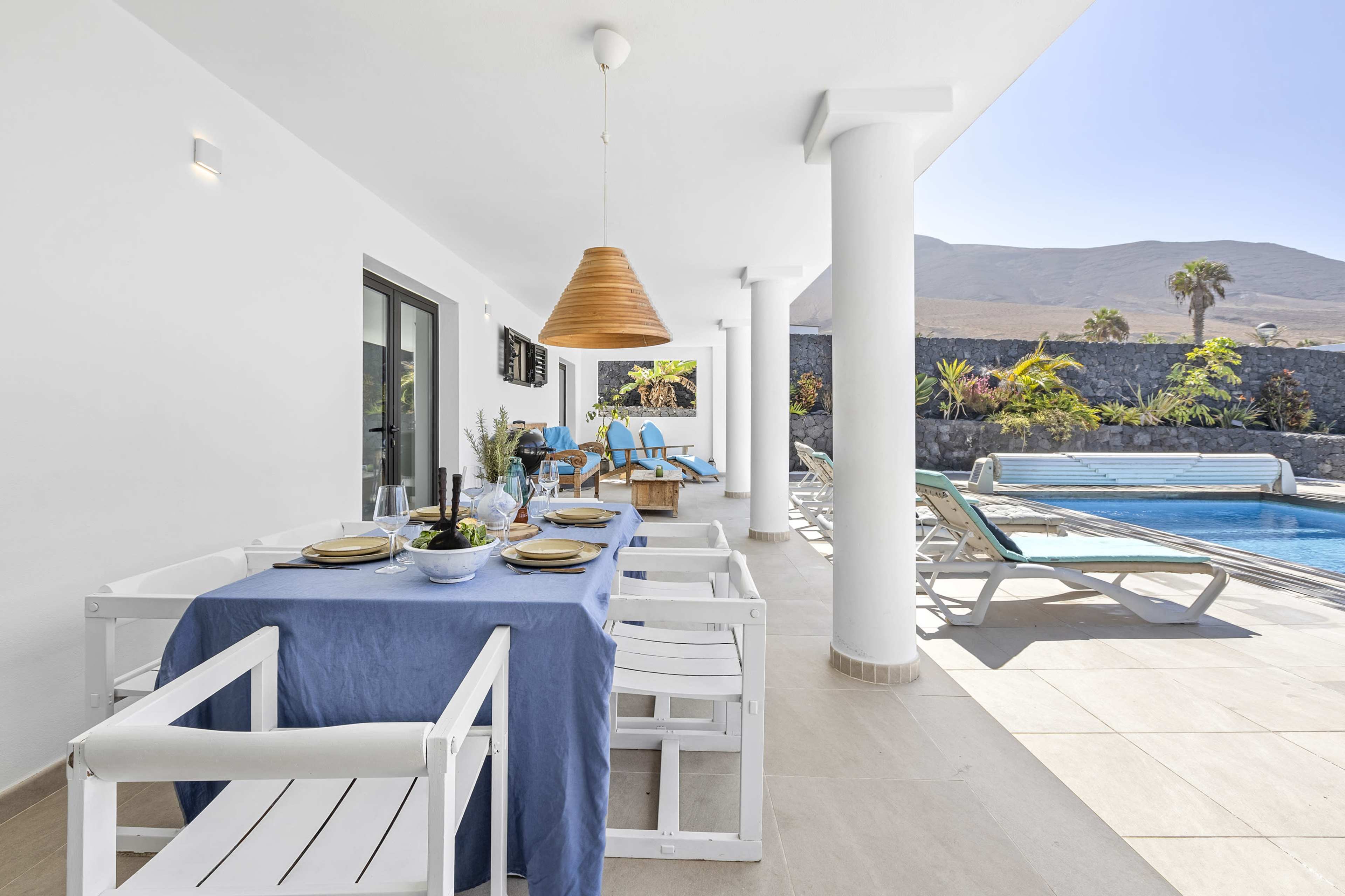 The image shows a modern outdoor patio area with a dining table set, lounge chairs, and a swimming pool surrounded by palm trees and mountains in the background.