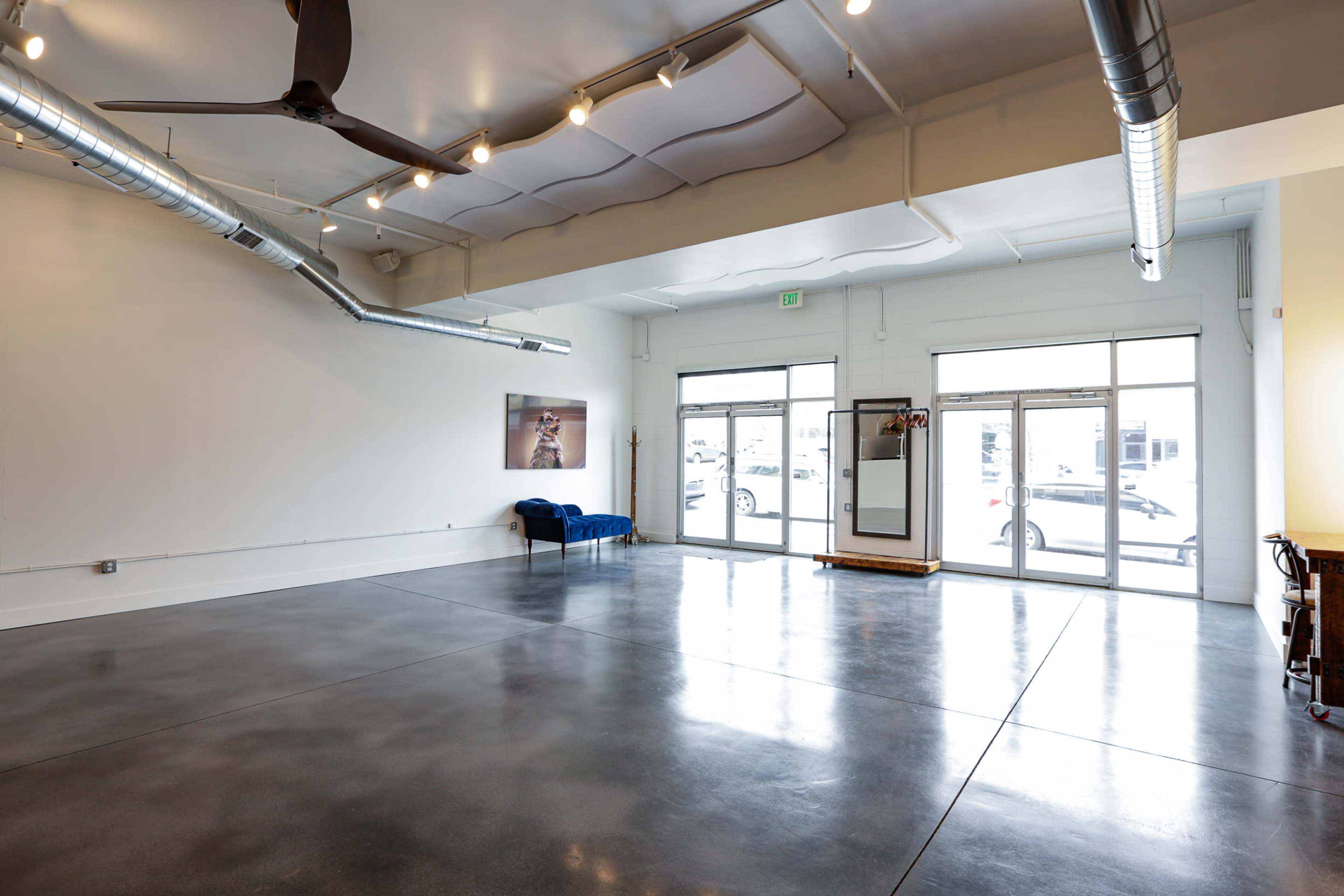 The image shows a spacious room with polished concrete floors, a blue couch against one wall, and large windows letting in natural light.