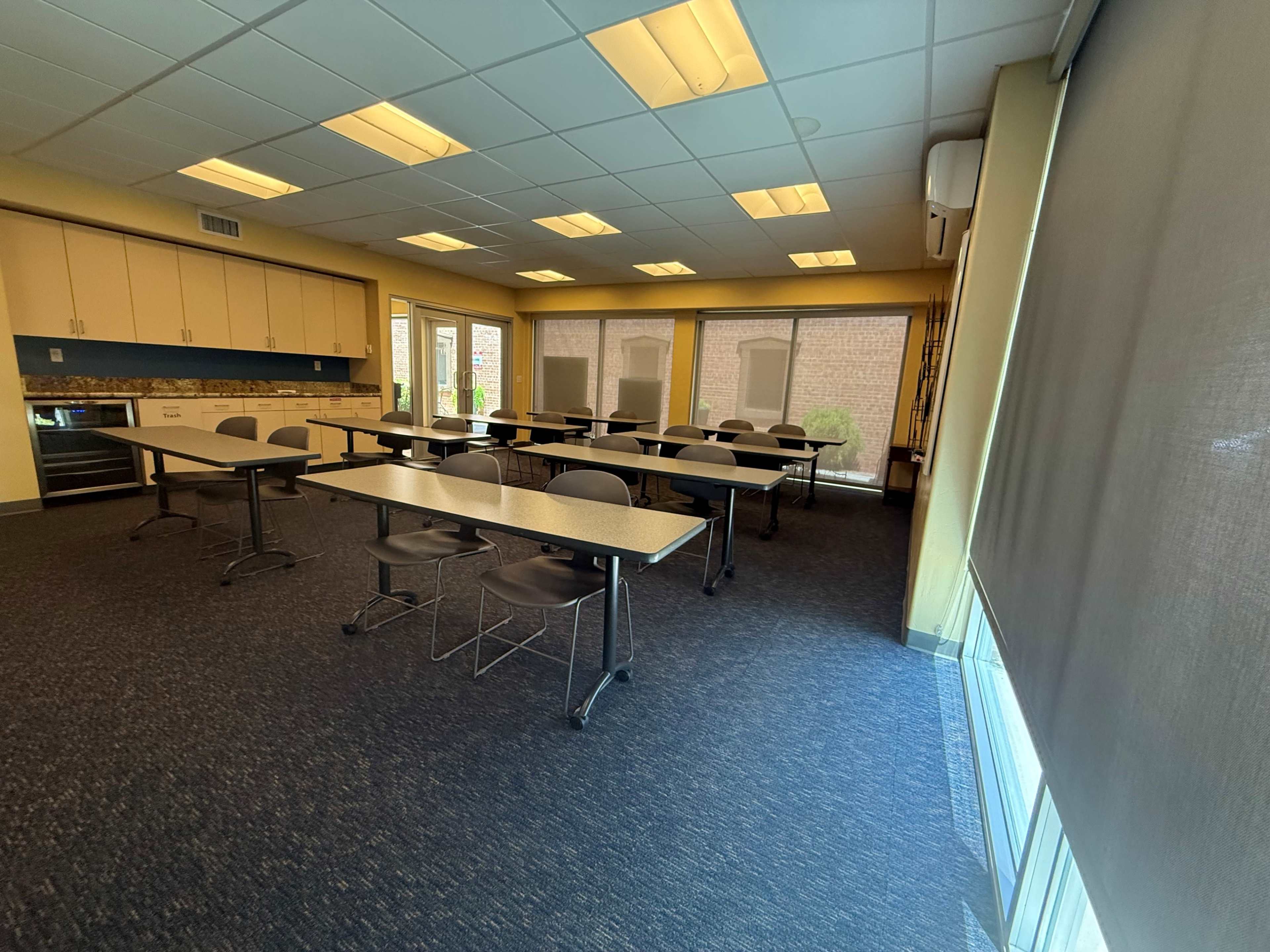 The image shows a classroom with several tables and chairs arranged in a spacious layout, featuring large windows and yellow walls.