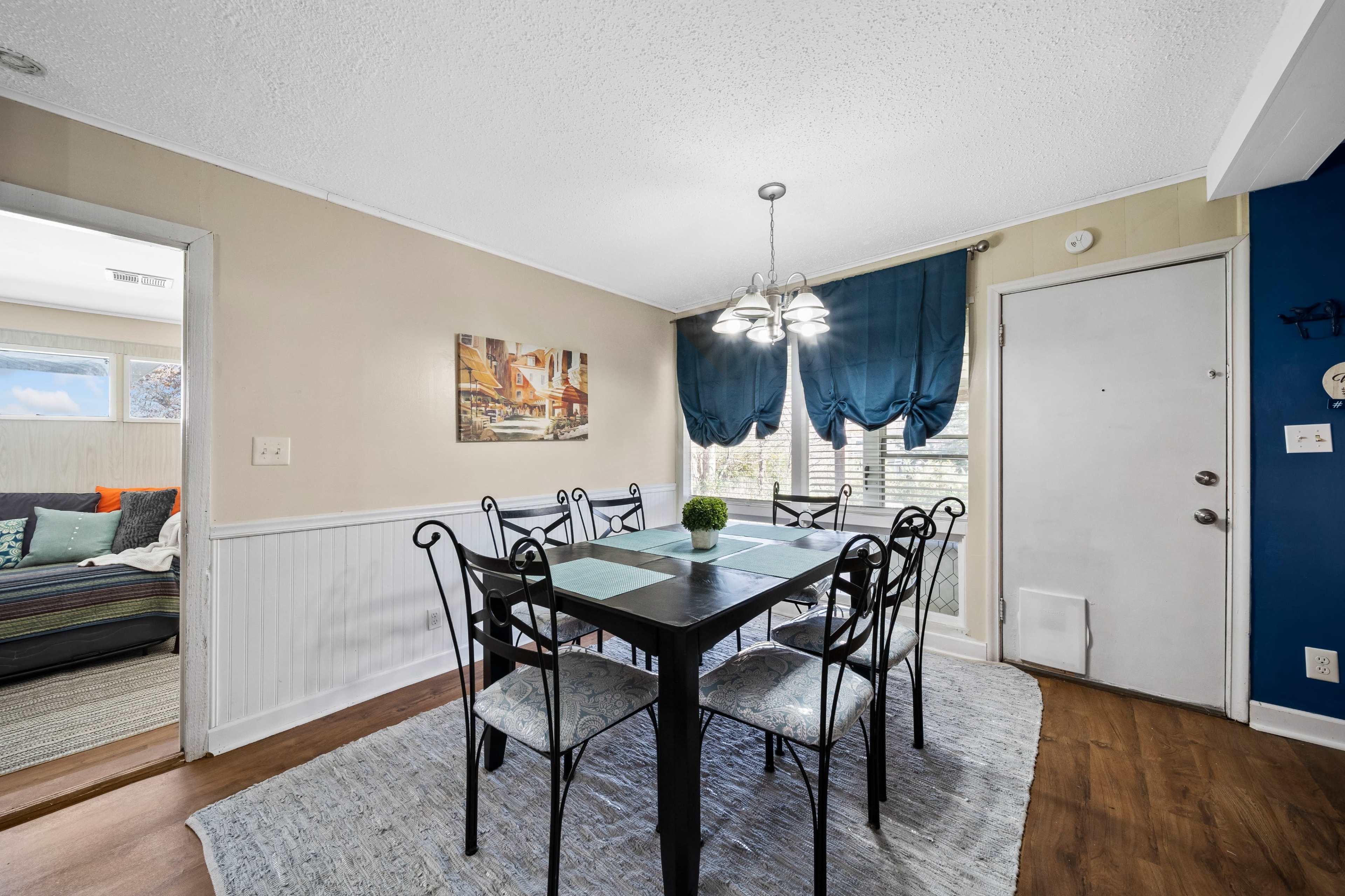 A dining area features a dark wooden table surrounded by six chairs, with blue drapes and a light fixture overhead.