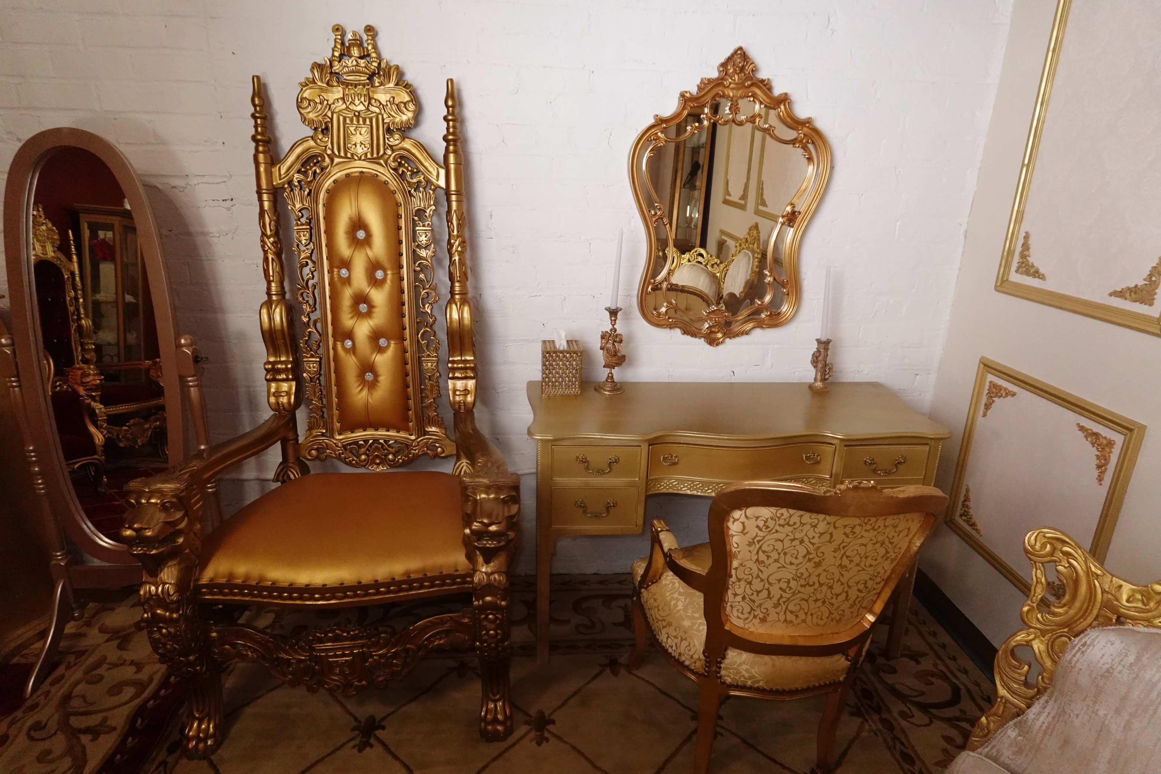 The image shows an ornate, golden throne chair beside a matching vanity table and a decorative mirror against a white brick wall.
