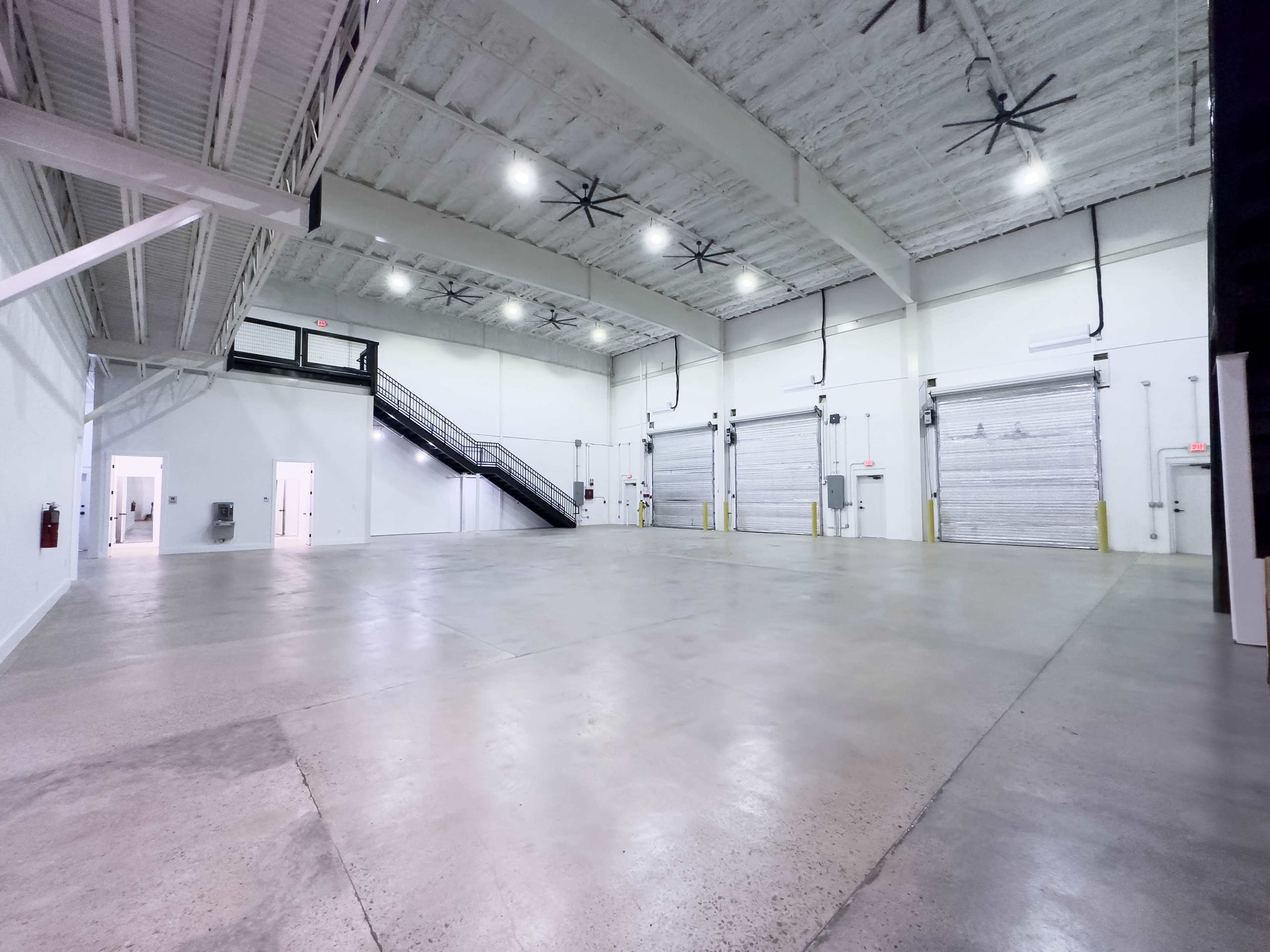 The image shows a spacious, empty warehouse with a high ceiling, large roll-up doors, and a staircase in the corner.