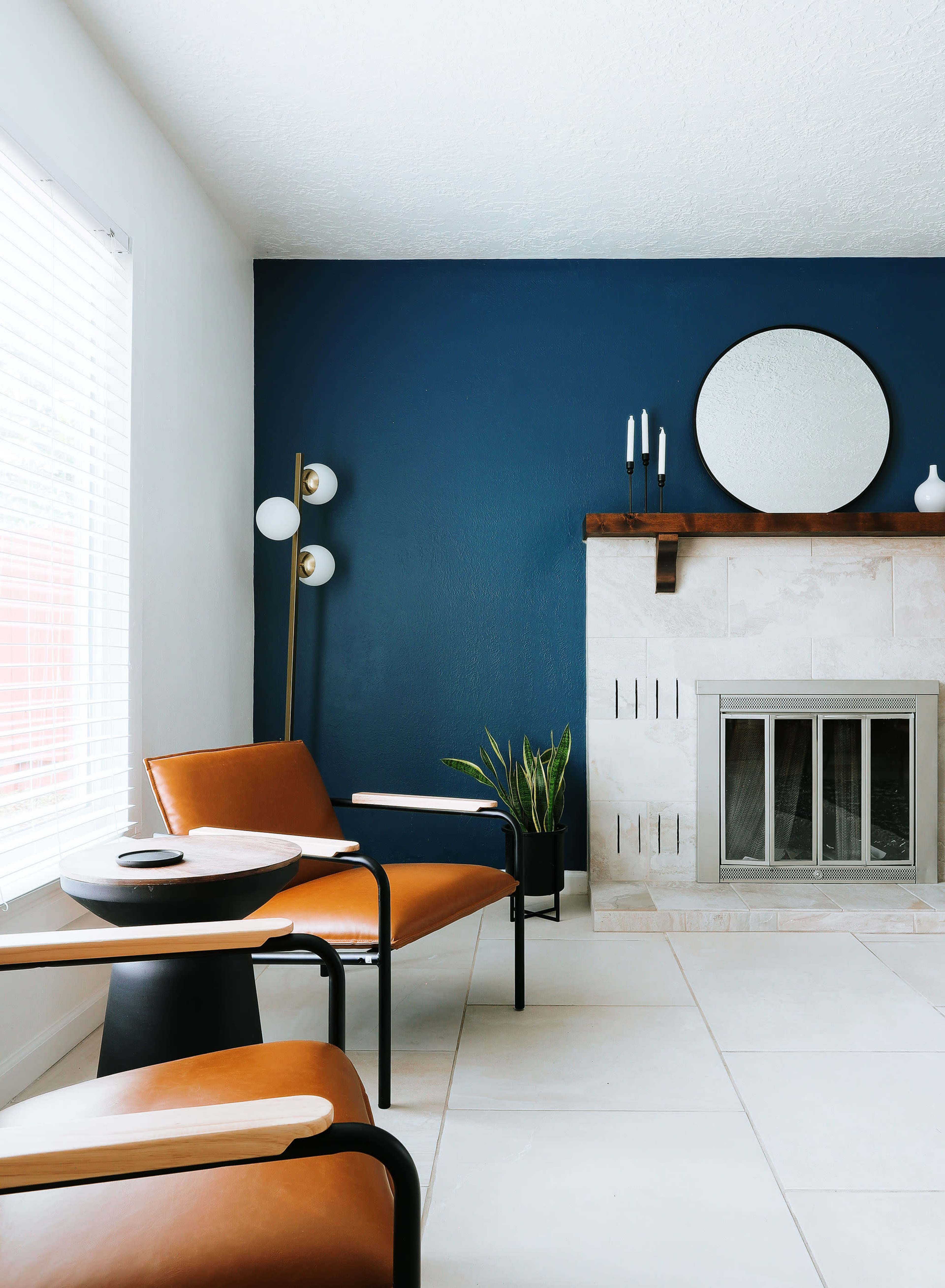 A modern living room with two brown leather chairs facing a stone fireplace and a circular mirror above it, accompanied by a small round table and a potted plant.