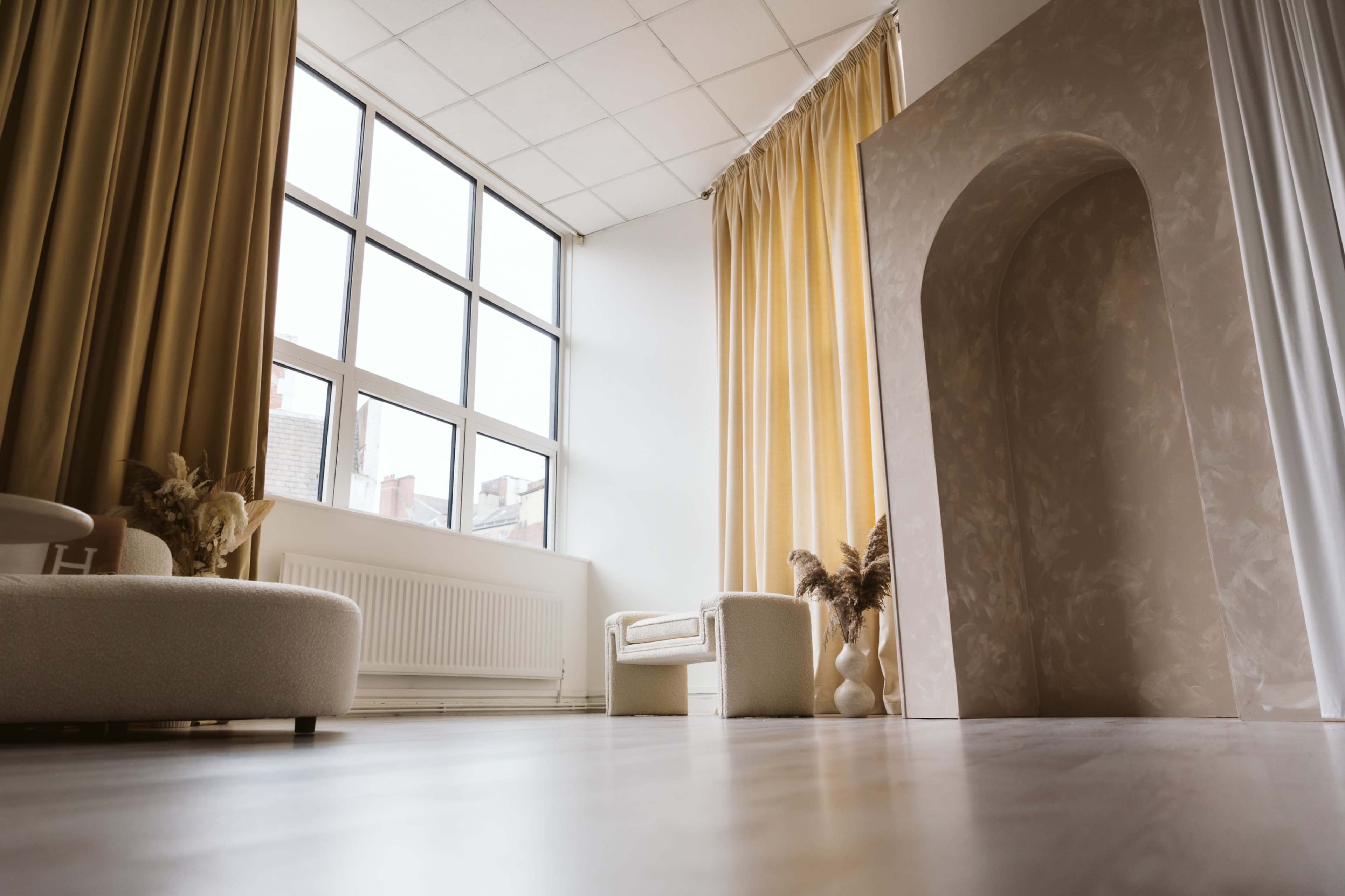 A spacious room with large windows, a light-colored archway, and minimal furniture including a sofa and an armchair.