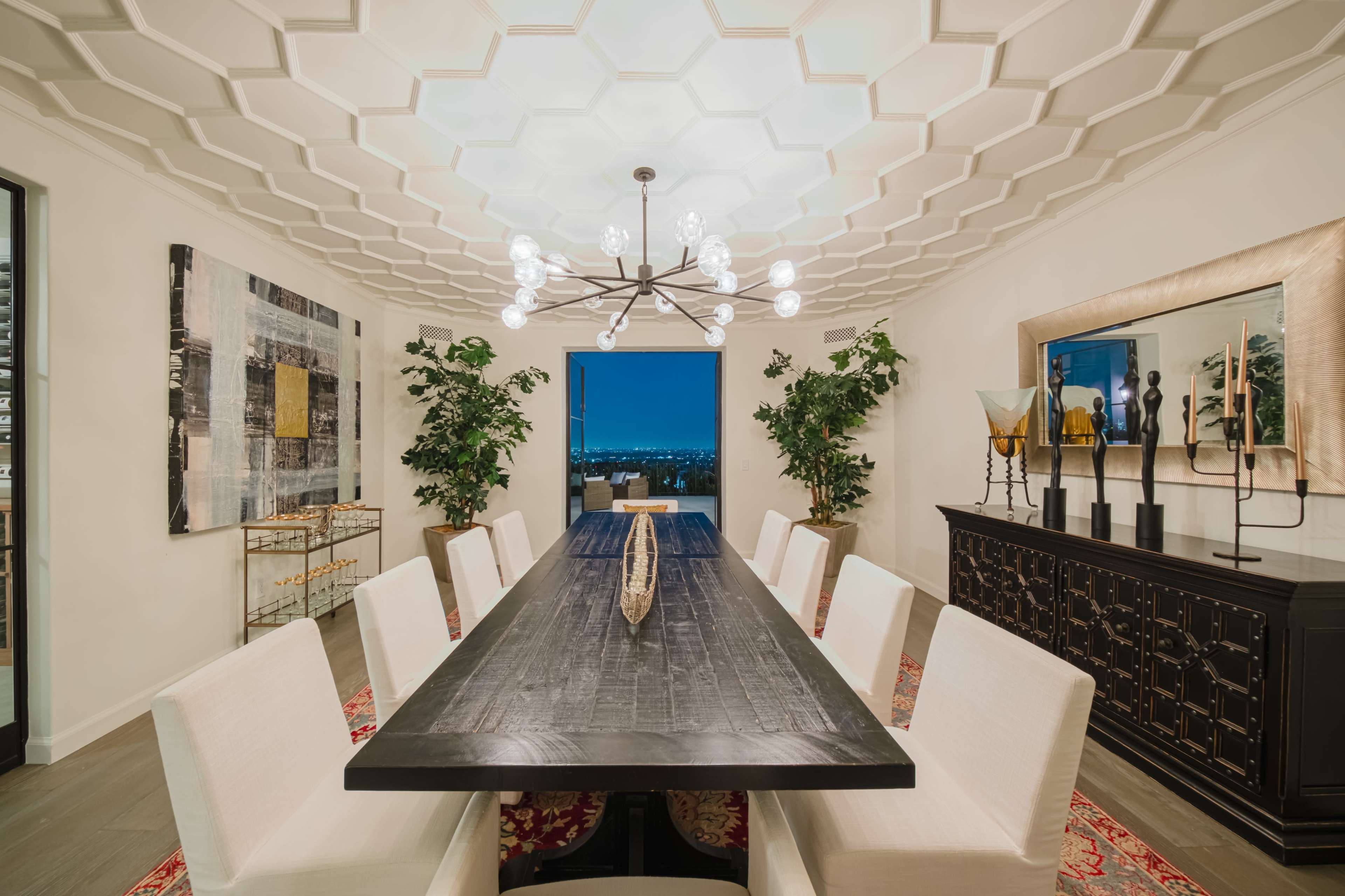 A modern dining room features a large wooden table surrounded by upholstered chairs, with a geometric ceiling design and large windows offering a view.