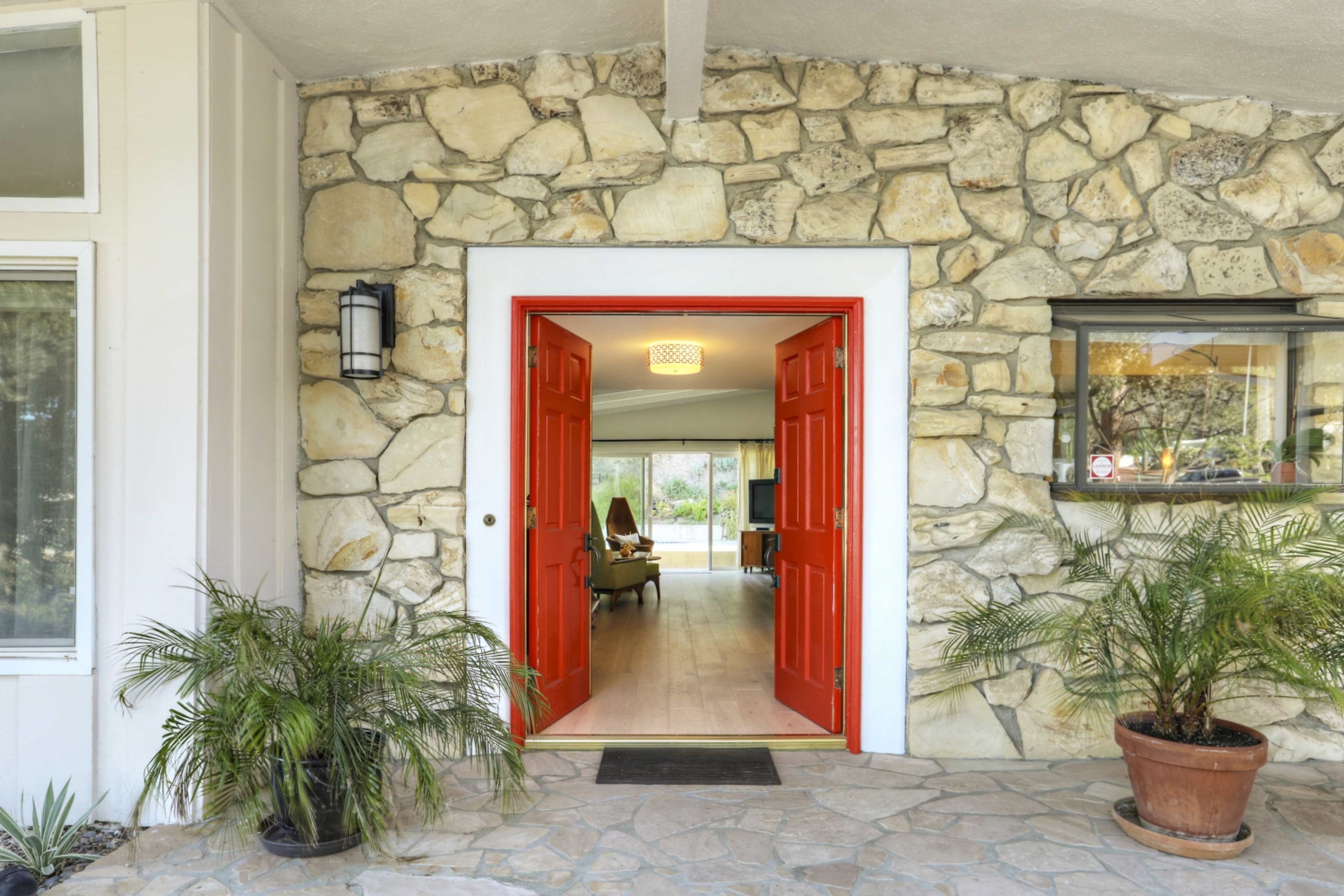A bright red double door framed by stone walls opens to a light-filled interior with a view of a spacious living area.