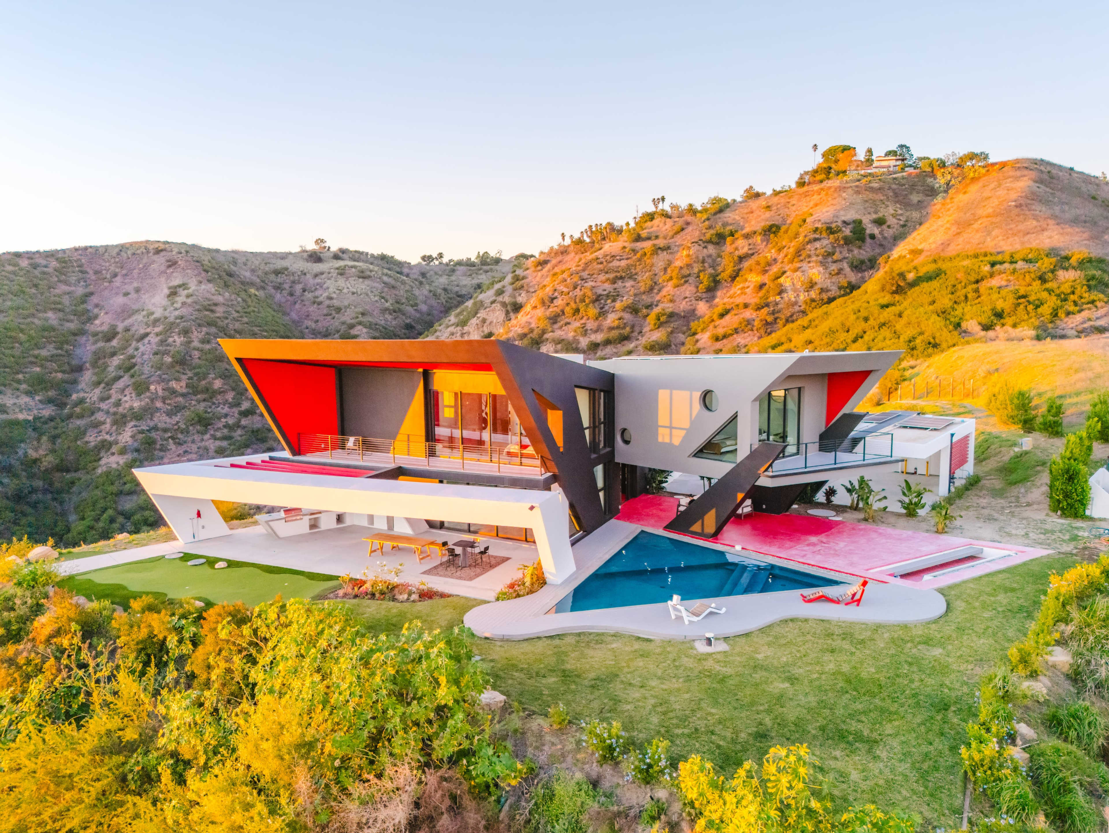 The image shows a modern house with angular architecture, featuring a red and black exterior, an outdoor pool, and surrounded by hilly terrain.