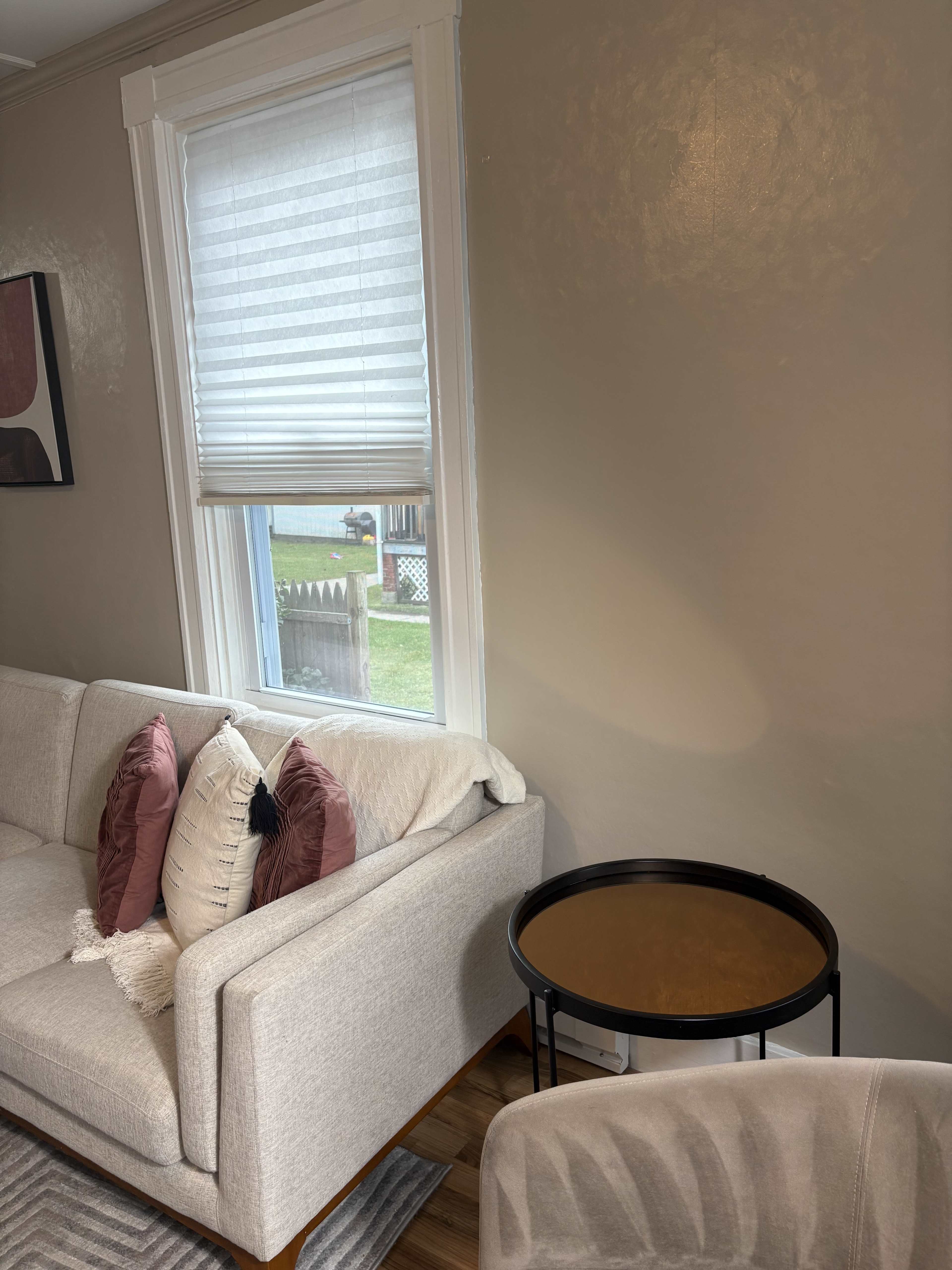 A light-colored sofa with decorative pillows is positioned near a window partially covered by a blind, next to a small black side table.
