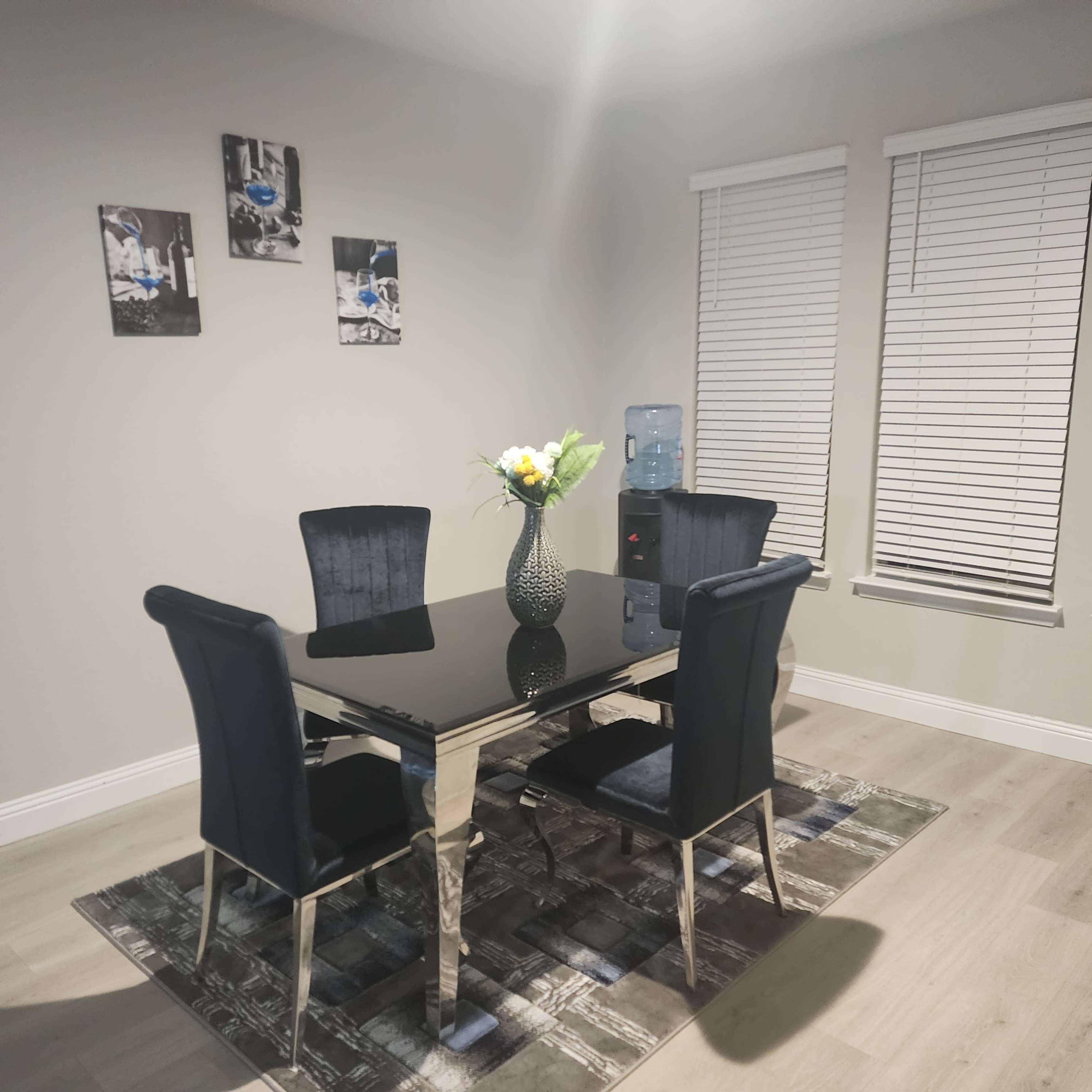 A glass-topped dining table with four black chairs is set on a patterned rug under two windows with blinds and framed artwork on the wall.