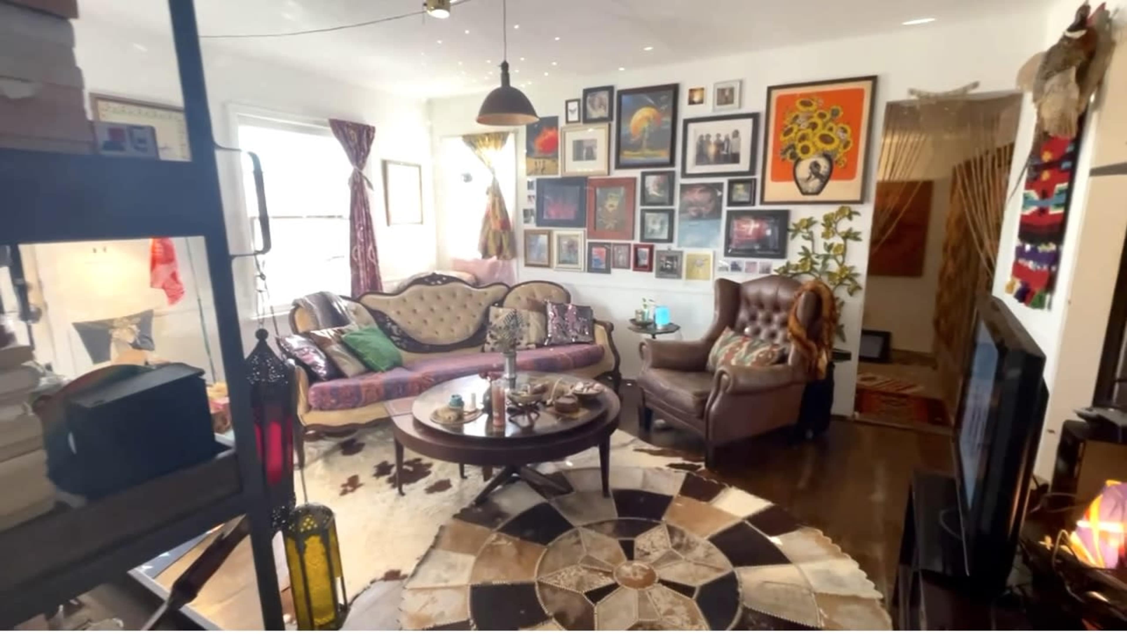 The room features a cozy seating area with a patterned rug, a round coffee table, and walls adorned with various framed artworks.