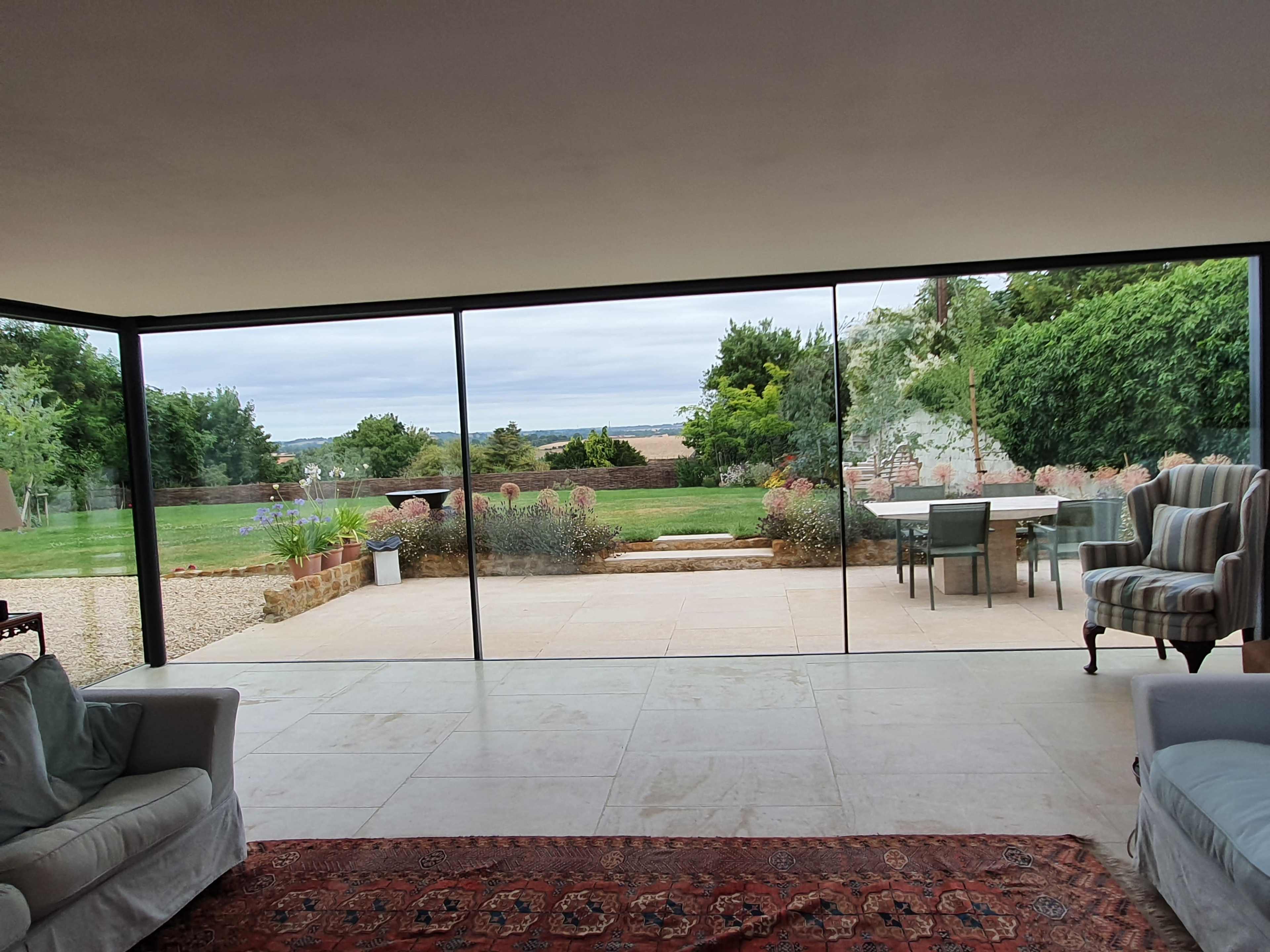 The image shows a spacious interior with large glass walls offering a view of a garden and sea in the distance.