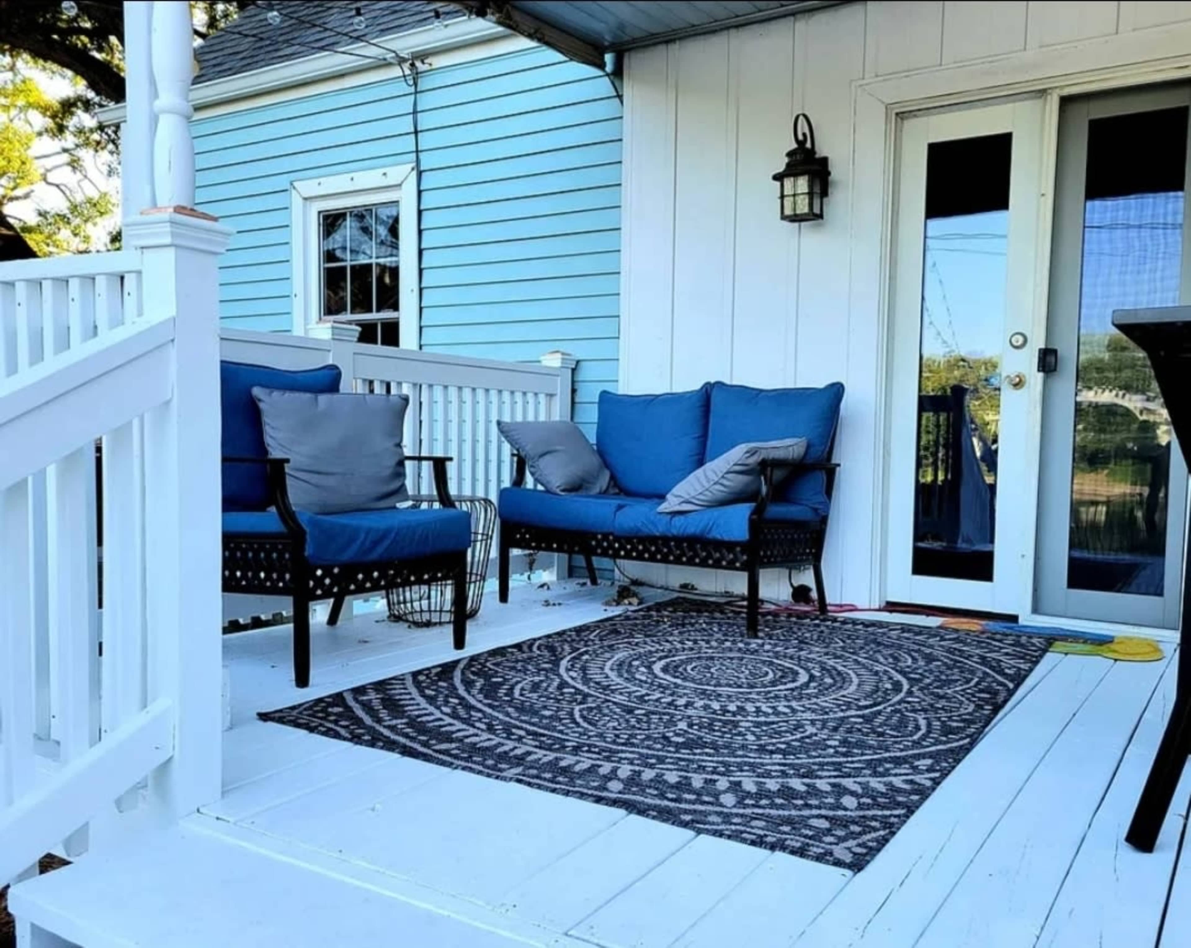 The image shows a porch area featuring two blue chairs on a patterned rug, with a white railing and a light blue house in the background.