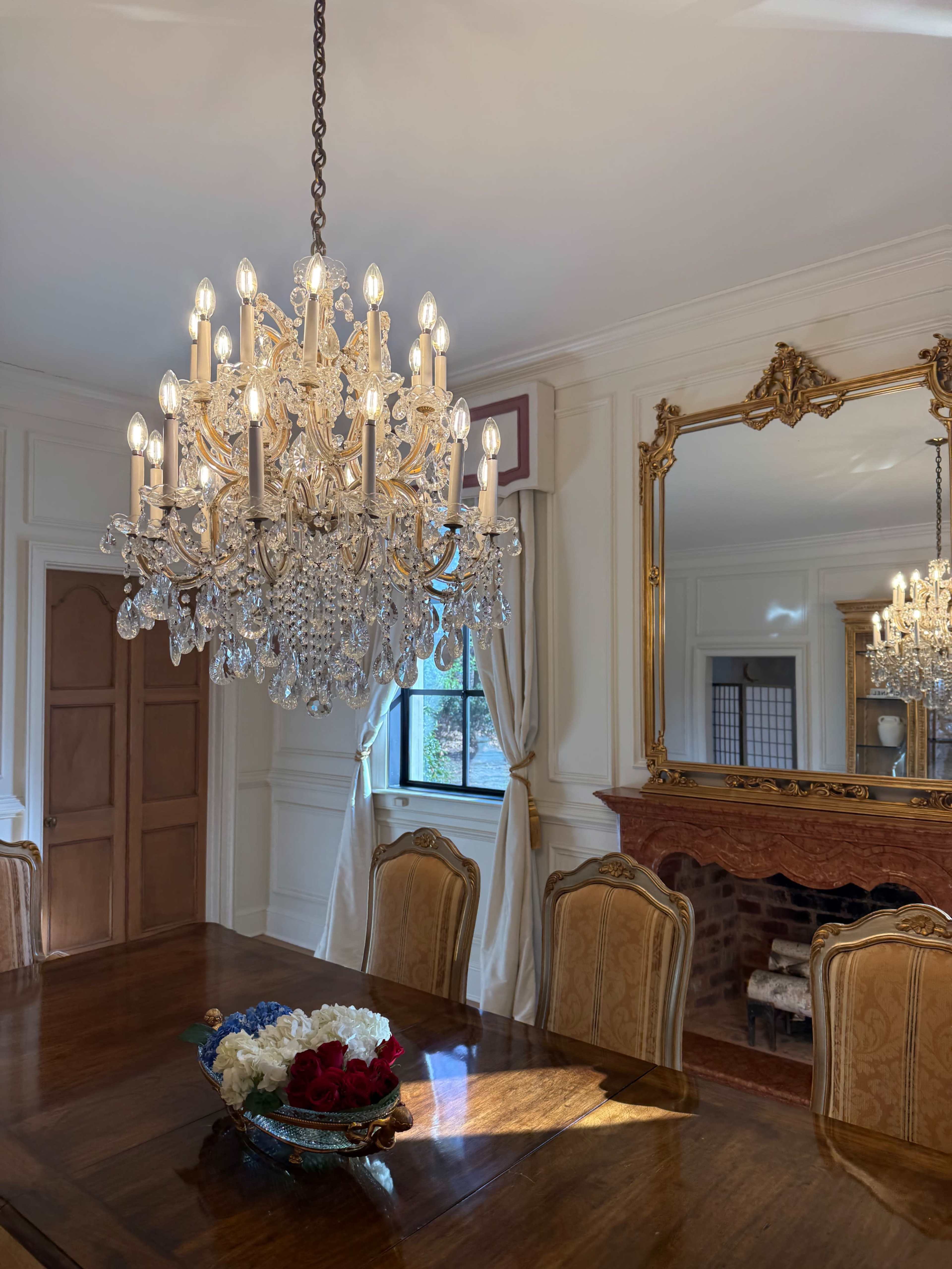 A large crystal chandelier hangs above a wooden dining table set with a decorative bowl of flowers, illuminated by natural light from a nearby window.