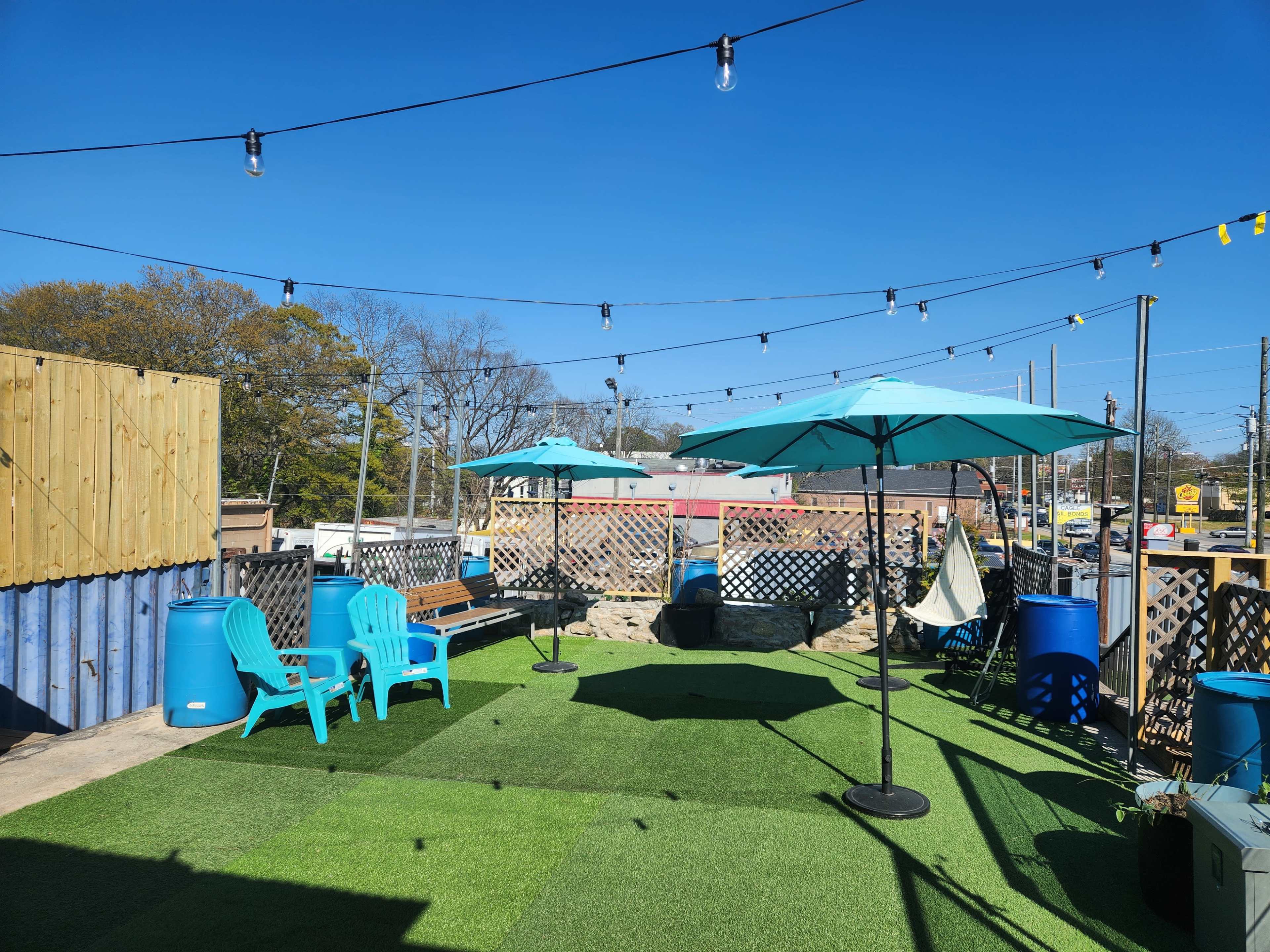 A rooftop patio area with green artificial grass, blue chairs, umbrellas, and string lights against a clear blue sky.